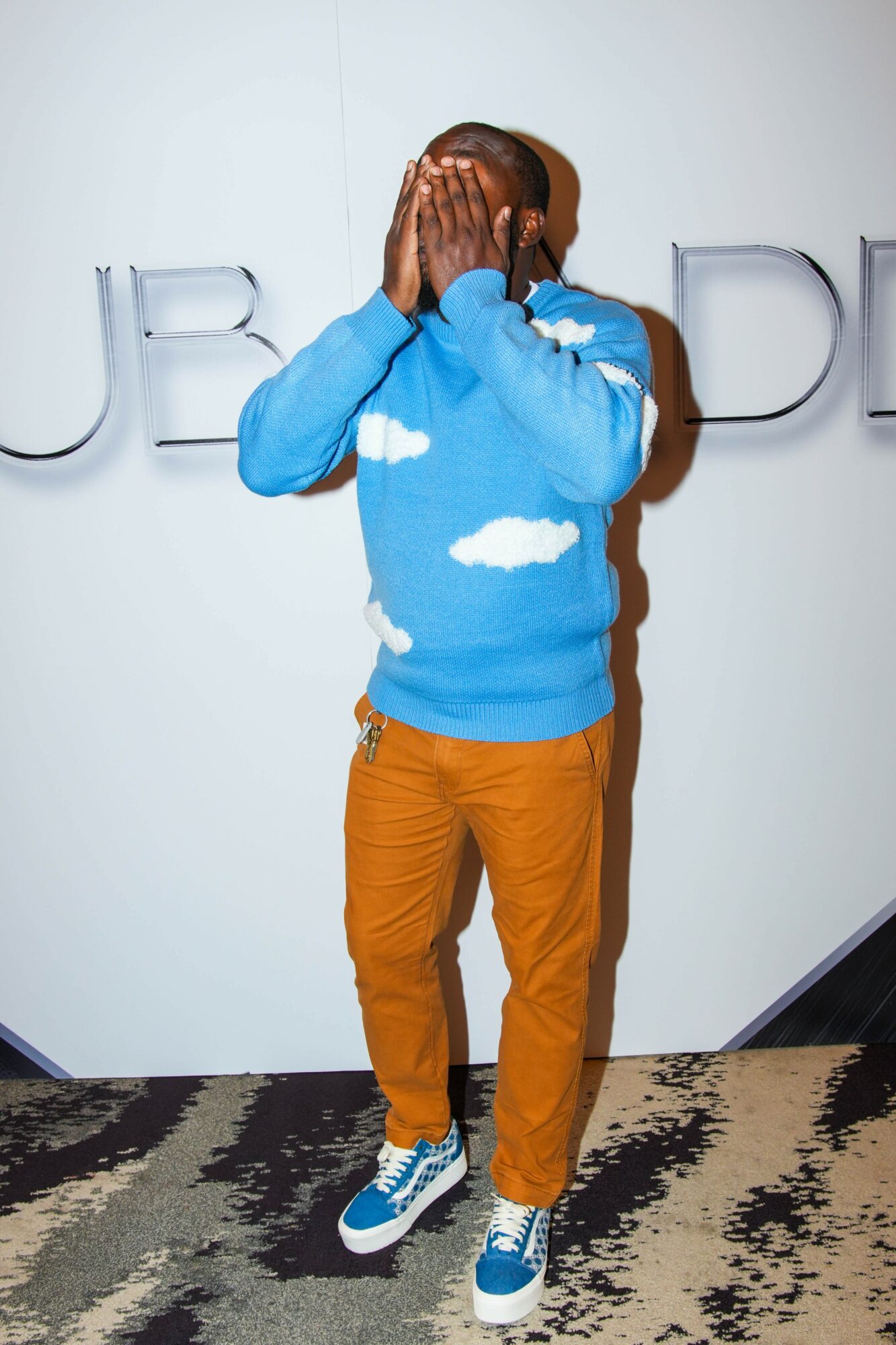 Person covering face with hands, wearing a blue cloud-patterned sweater and orange pants, standing on a patterned carpet.