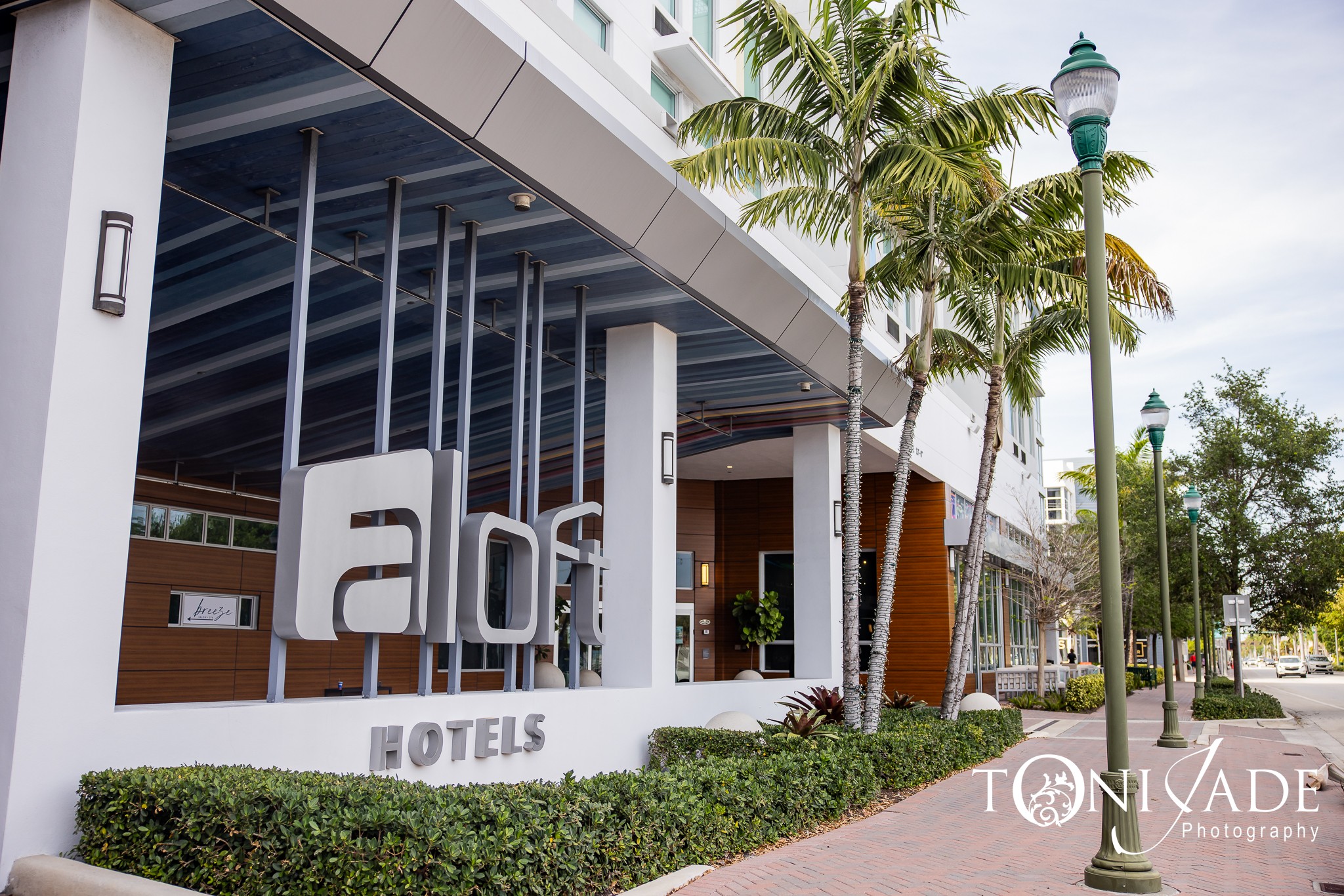 Hotel entrance with large sign, palm trees, and a sidewalk with street lamps, under a partly cloudy sky.