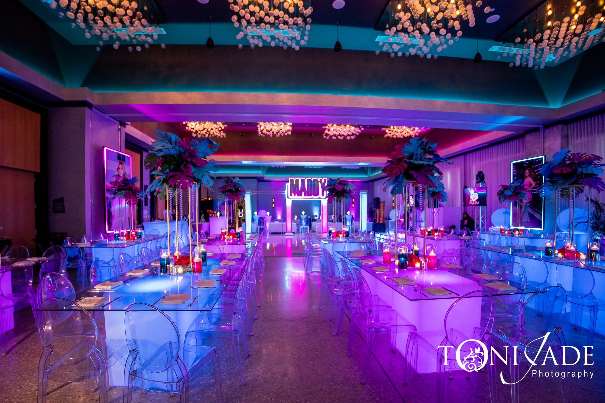 Elegant event space decorated with colorful lighting, tables, and floral arrangements, with a stage at the far end.