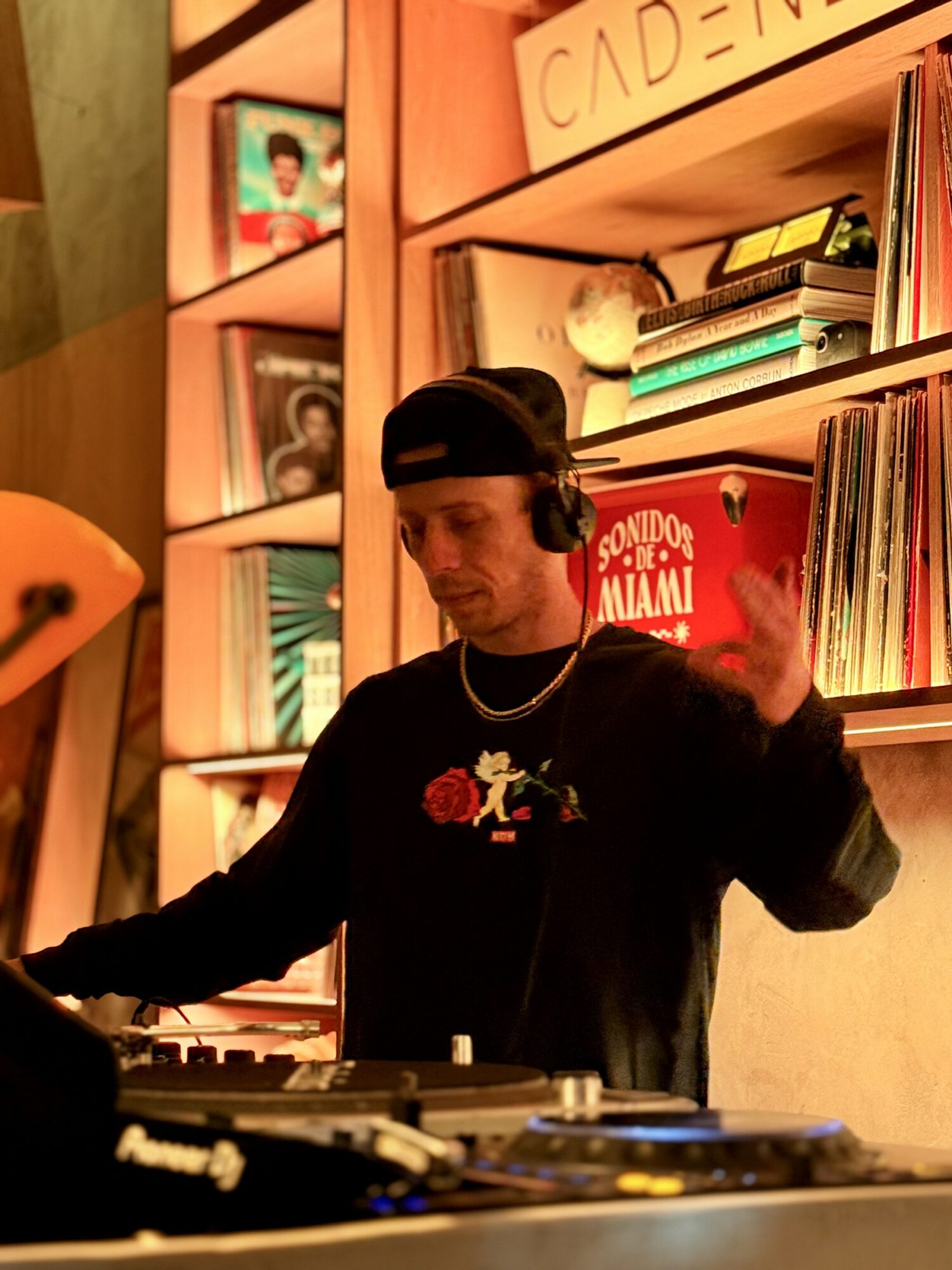 Young man wearing a black cap and sweatshirt with a DJ setup, shelves with books and records in background.