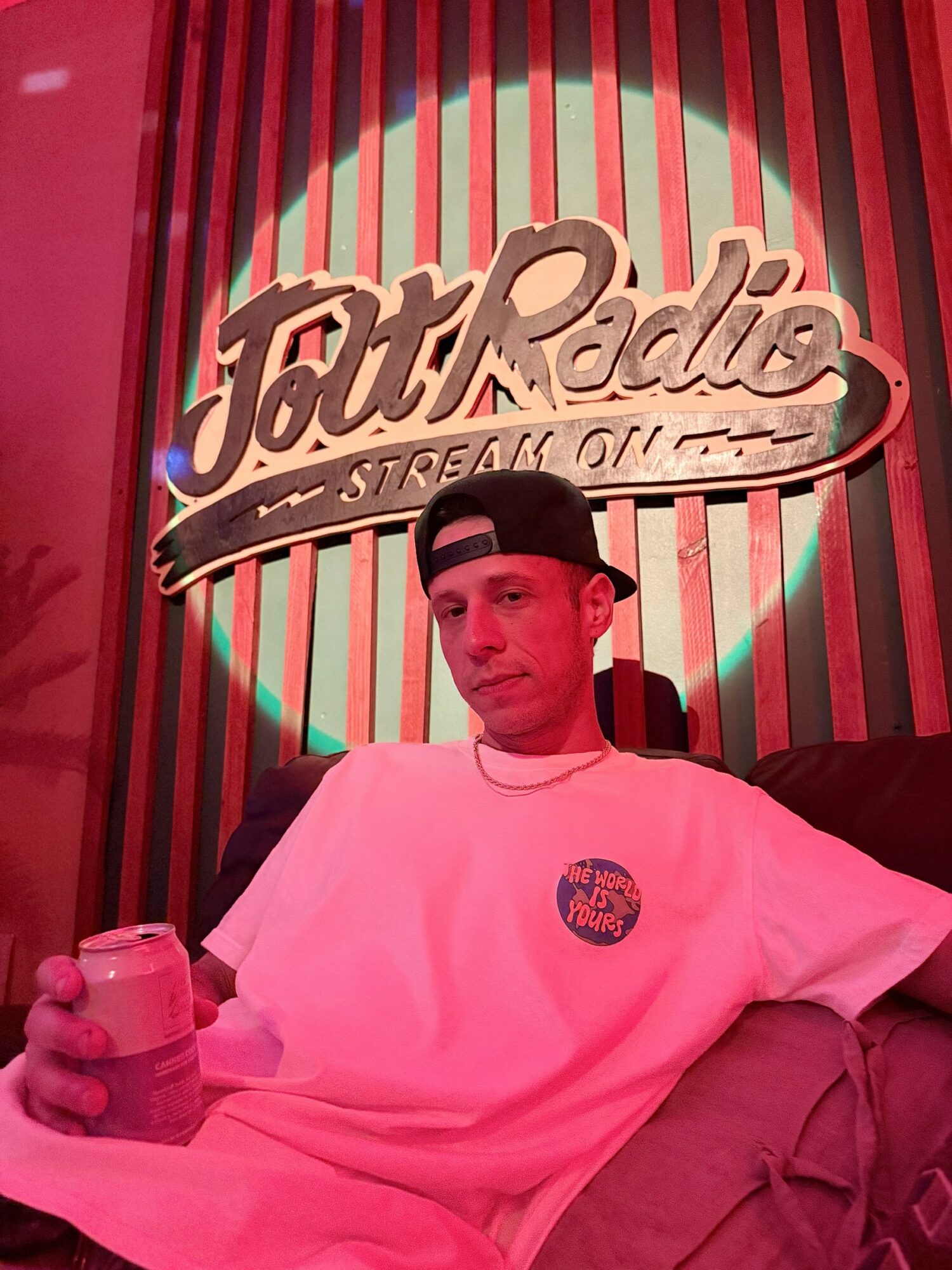 Young man in white shirt and black cap sitting in front of a TouRado sign, holding a drink, with red and green background.
