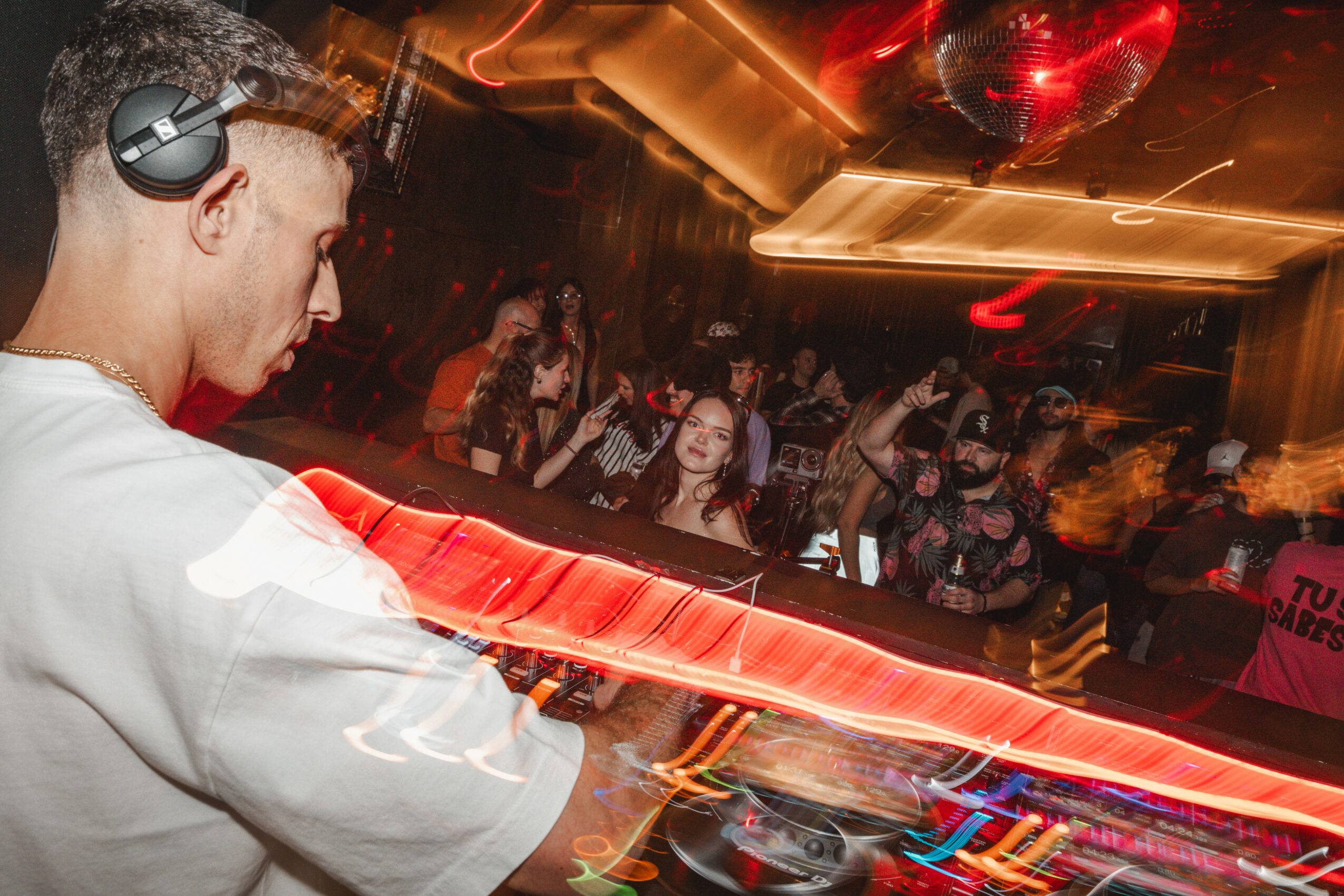 Person wearing headphones in a nightclub with colorful lights and a crowd dancing in the background.