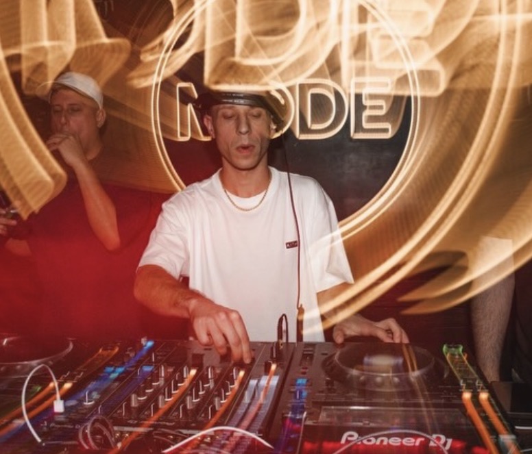 DJ wearing headphones and a white shirt, mixing music at a club with colorful lights and a Pioneer DJ controller.