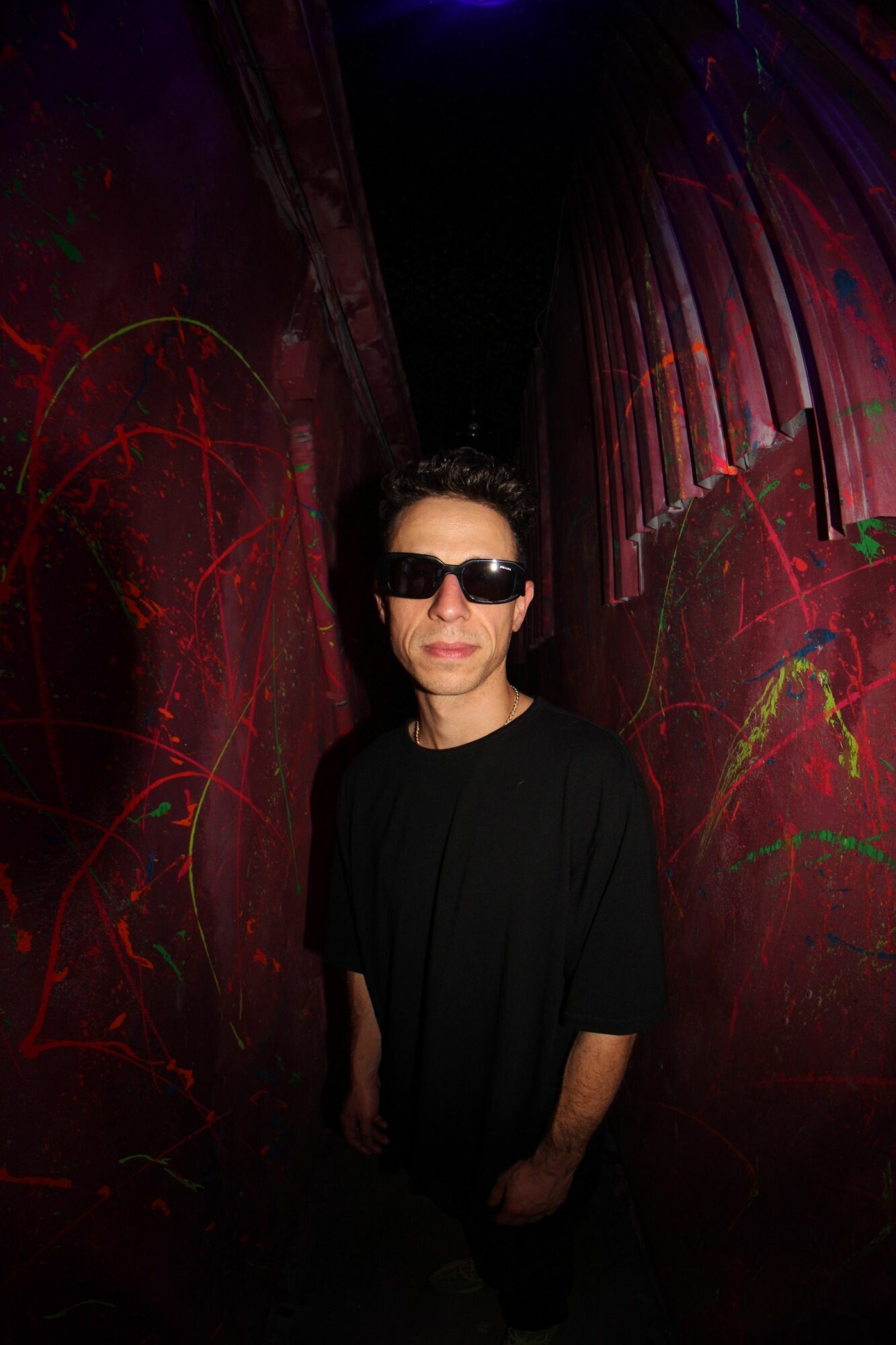 Person wearing sunglasses and black clothing standing in a dark, colorful, abstract environment.