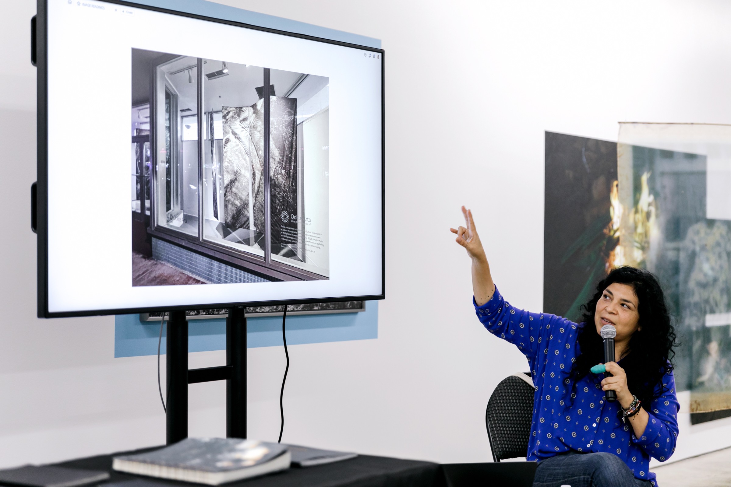 Woman presenting a slide of a glass shower enclosure on a large screen, sitting in a room with white walls.