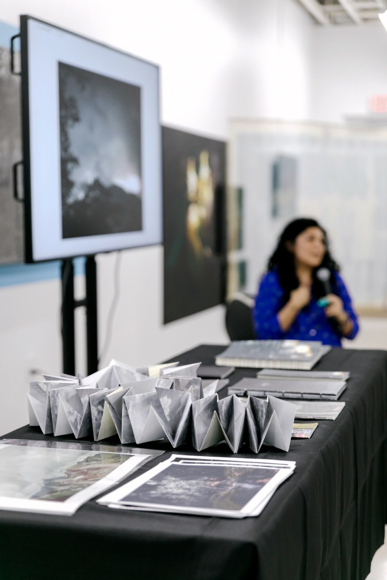 Table with folded paper objects and photographs, blurred woman in blue shirt in background, art gallery setting.