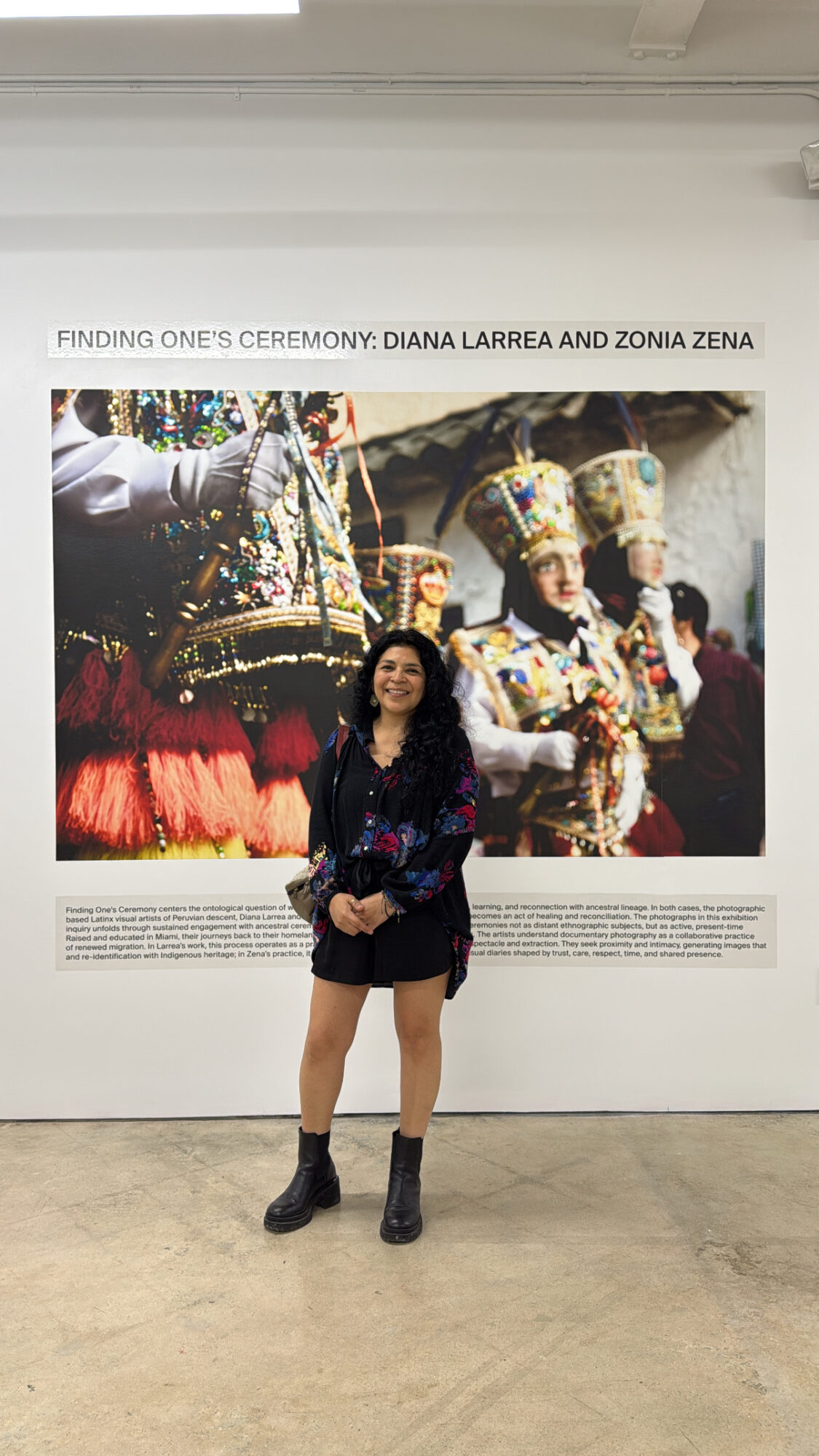 Person standing in front of an art display with traditional costumes, smiling, wearing a dark jacket, shorts, and boots.