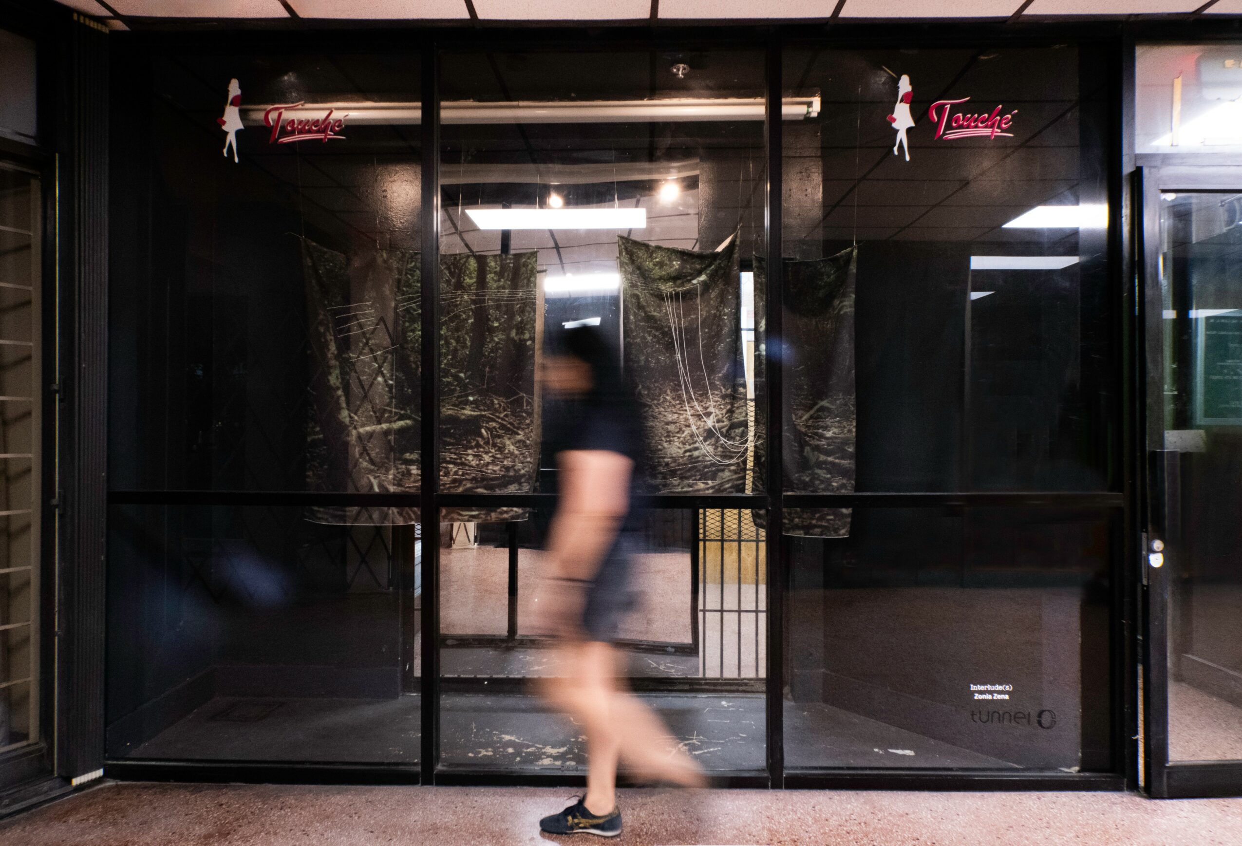 Person walking past a glass storefront with curtains inside, reflections visible on the glass, and pink signs hanging above.