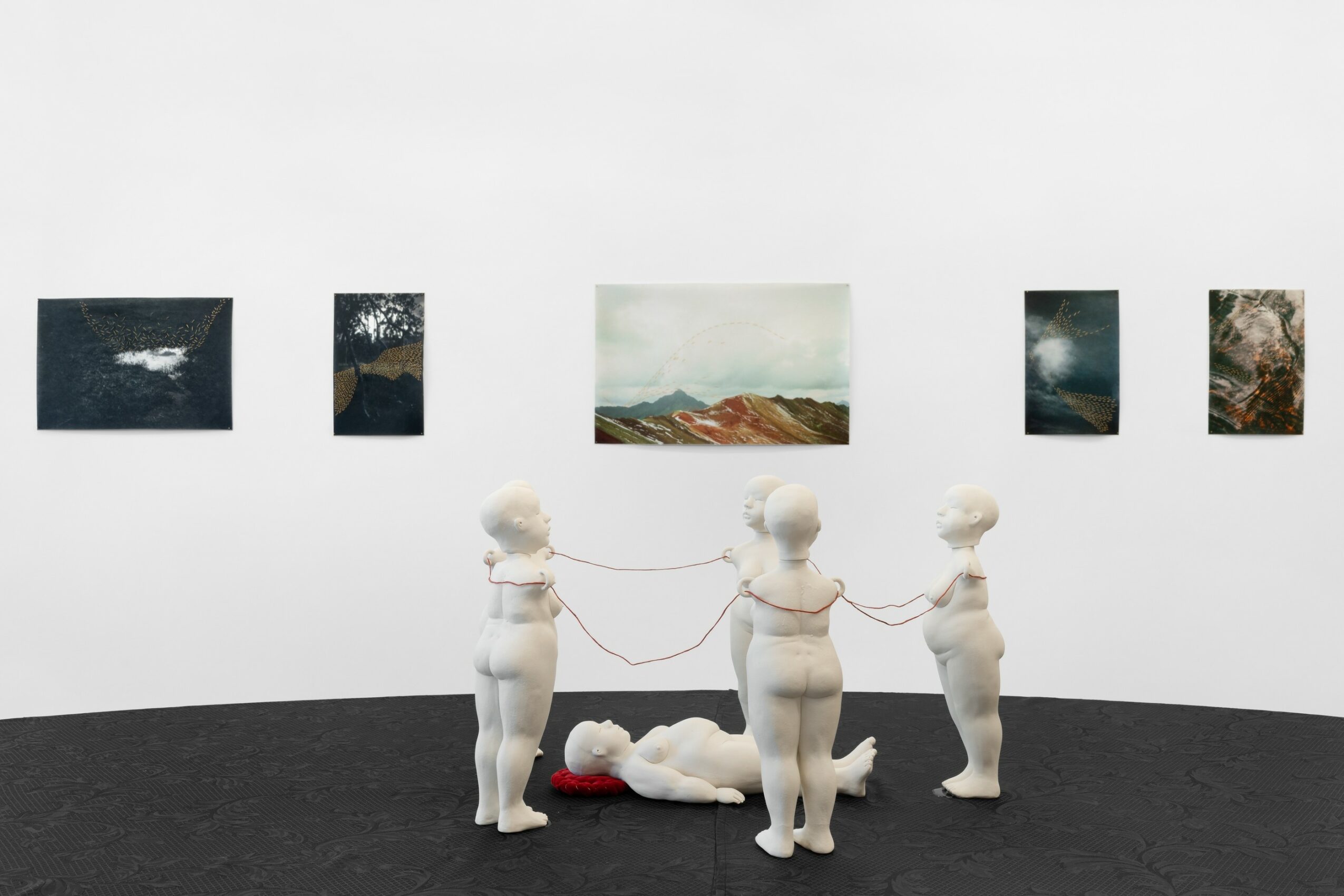 Four sculptures of children holding a string, with a child lying on the floor, in front of landscape paintings on a white wall.