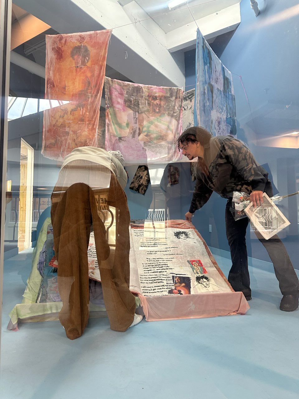 Two people are looking at an exhibit with informational panels in a gallery. One person is holding a newspaper, and the other is wearing a white headscarf. The exhibit includes colorful artwork and text panels.