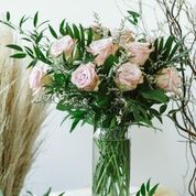 A bouquet of pale pink roses with green leaves in a tall glass vase, set against a white background.