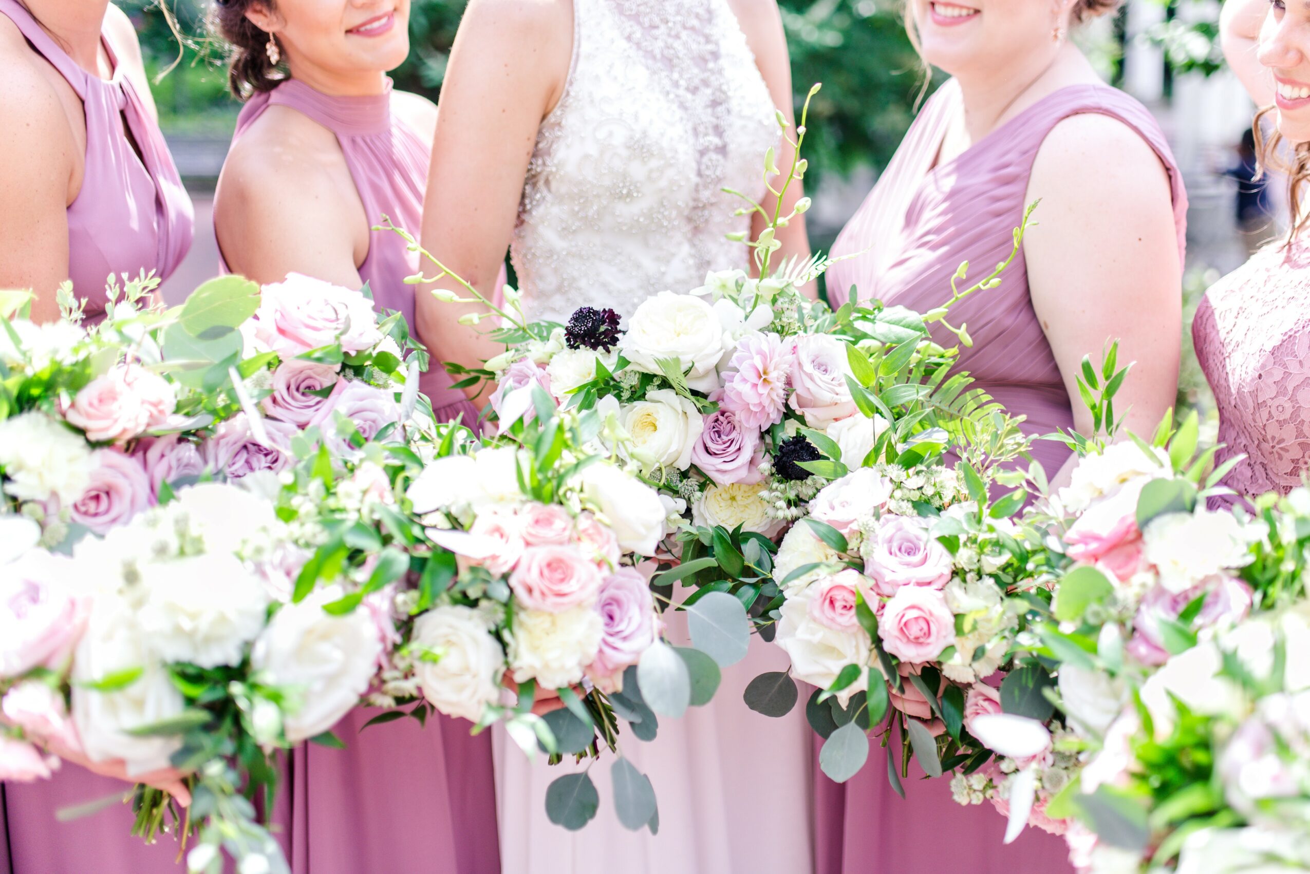 Group of women in pink dresses holding bouquets of pink and white flowers at a wedding, with greenery in background.