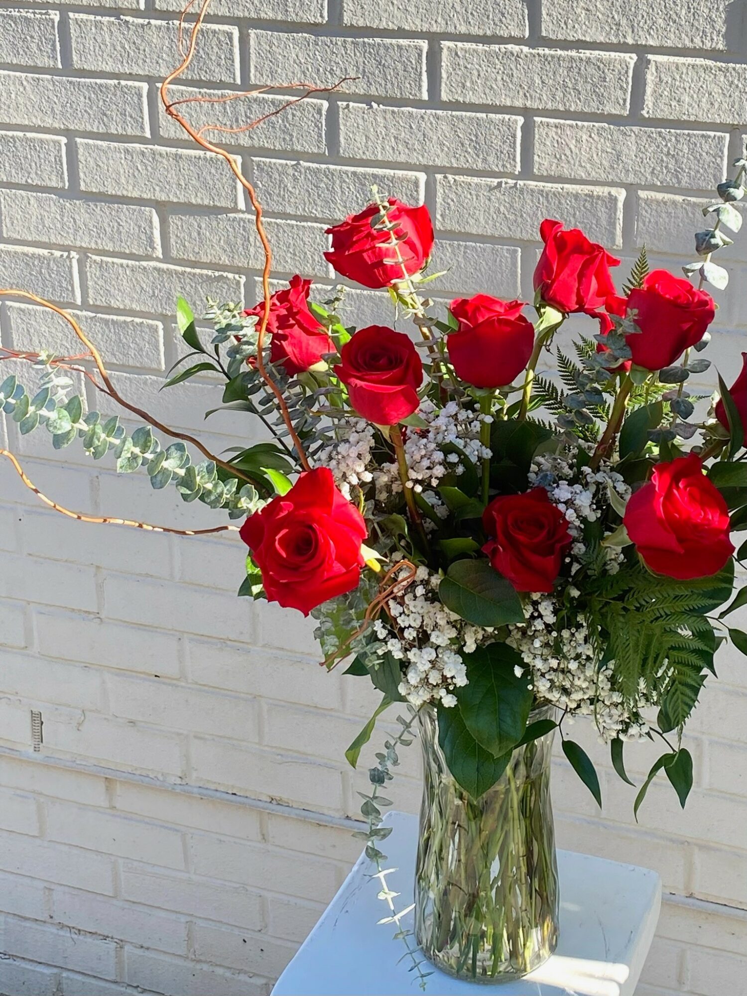 Vase with red roses, white baby's breath, and green leaves against a brick wall, on a white surface.