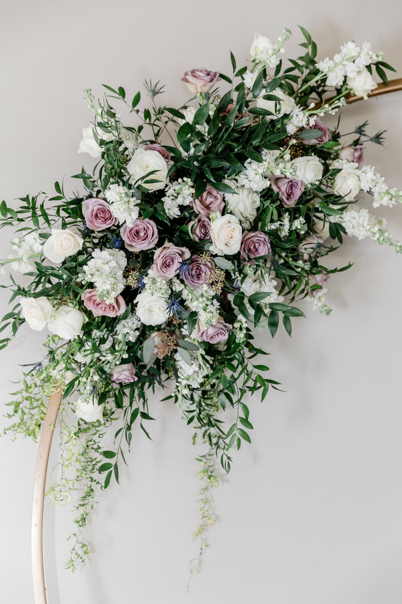 A cascading floral arrangement with white and purple roses, white hydrangeas, and green foliage against a plain background.