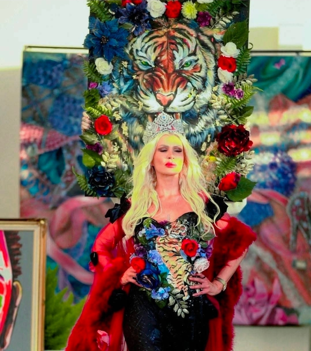 Woman with long blonde hair in front of a tiger and floral backdrop, holding a bouquet of flowers, wearing a black dress with floral details.