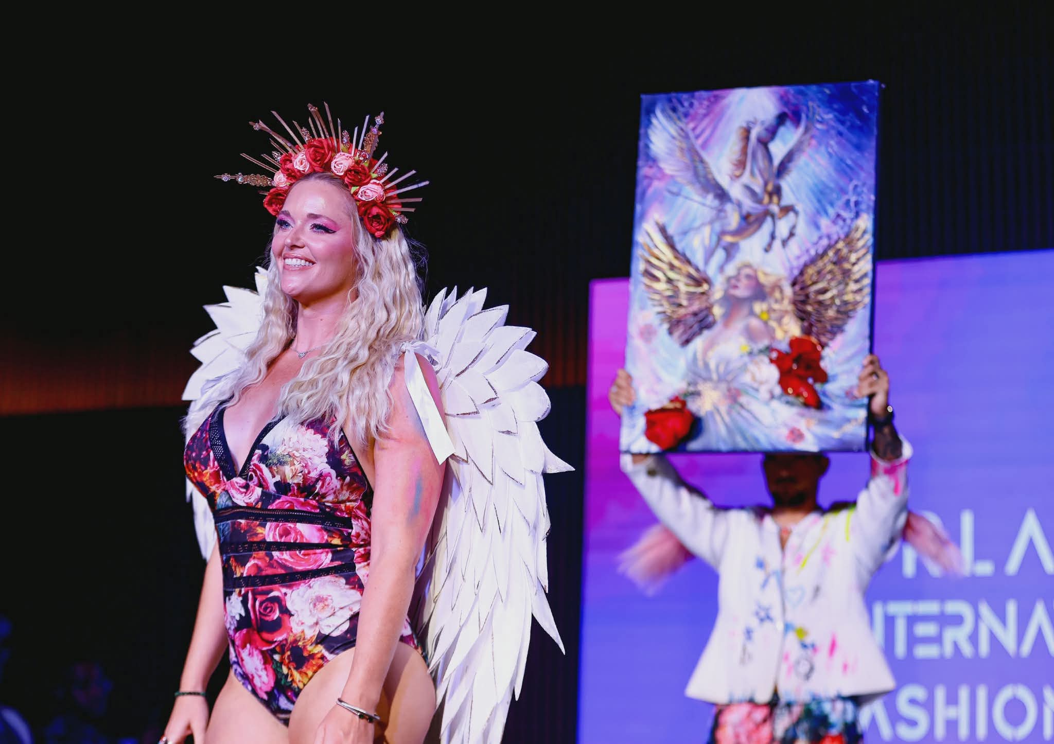 Woman with a floral crown and feathered costume on stage, smiling, with a person holding a colorful painting in the background.