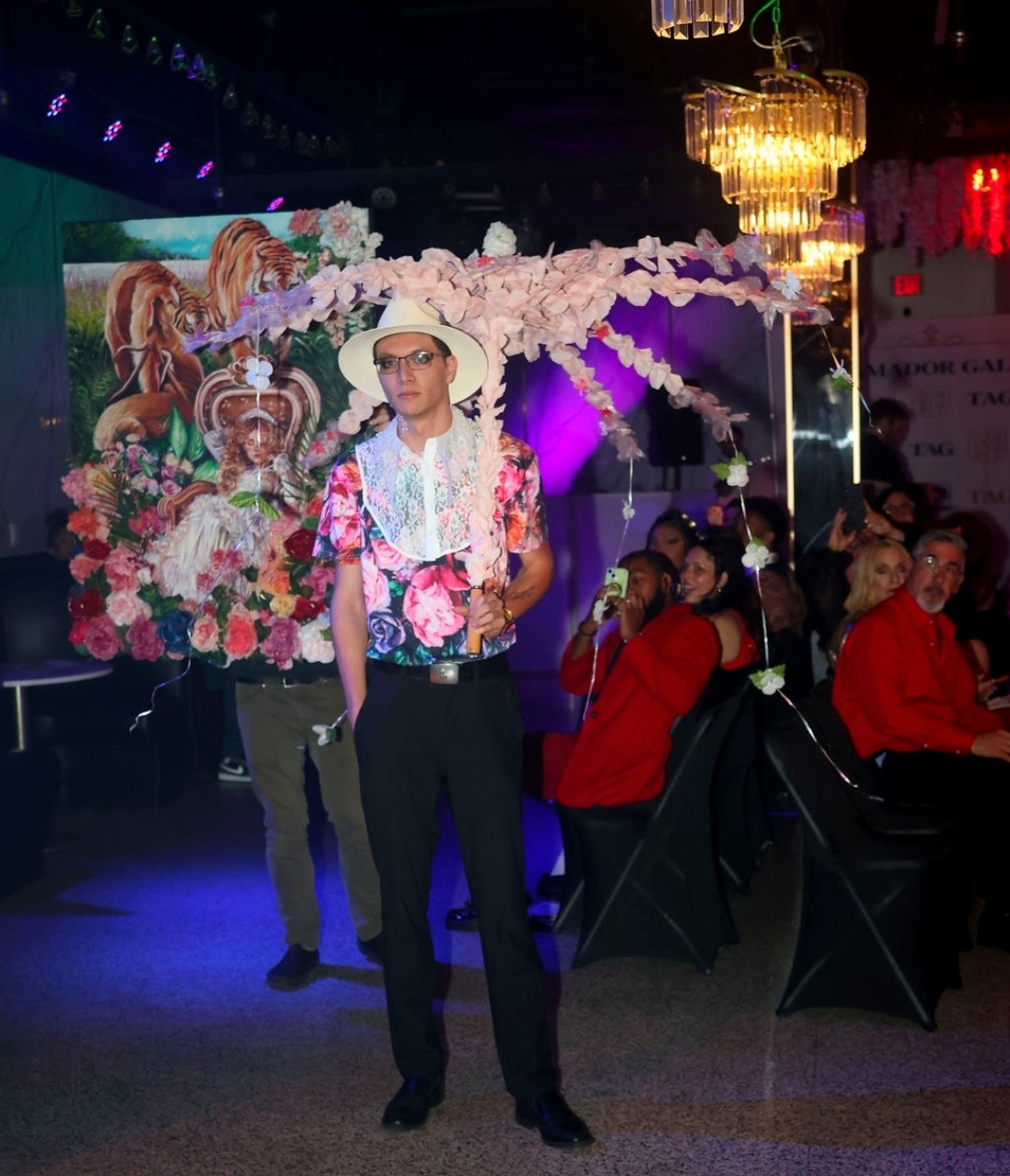 Person walking on runway with floral and animal-themed backdrop, wearing a hat, sunglasses, and colorful shirt, audience seated nearby.