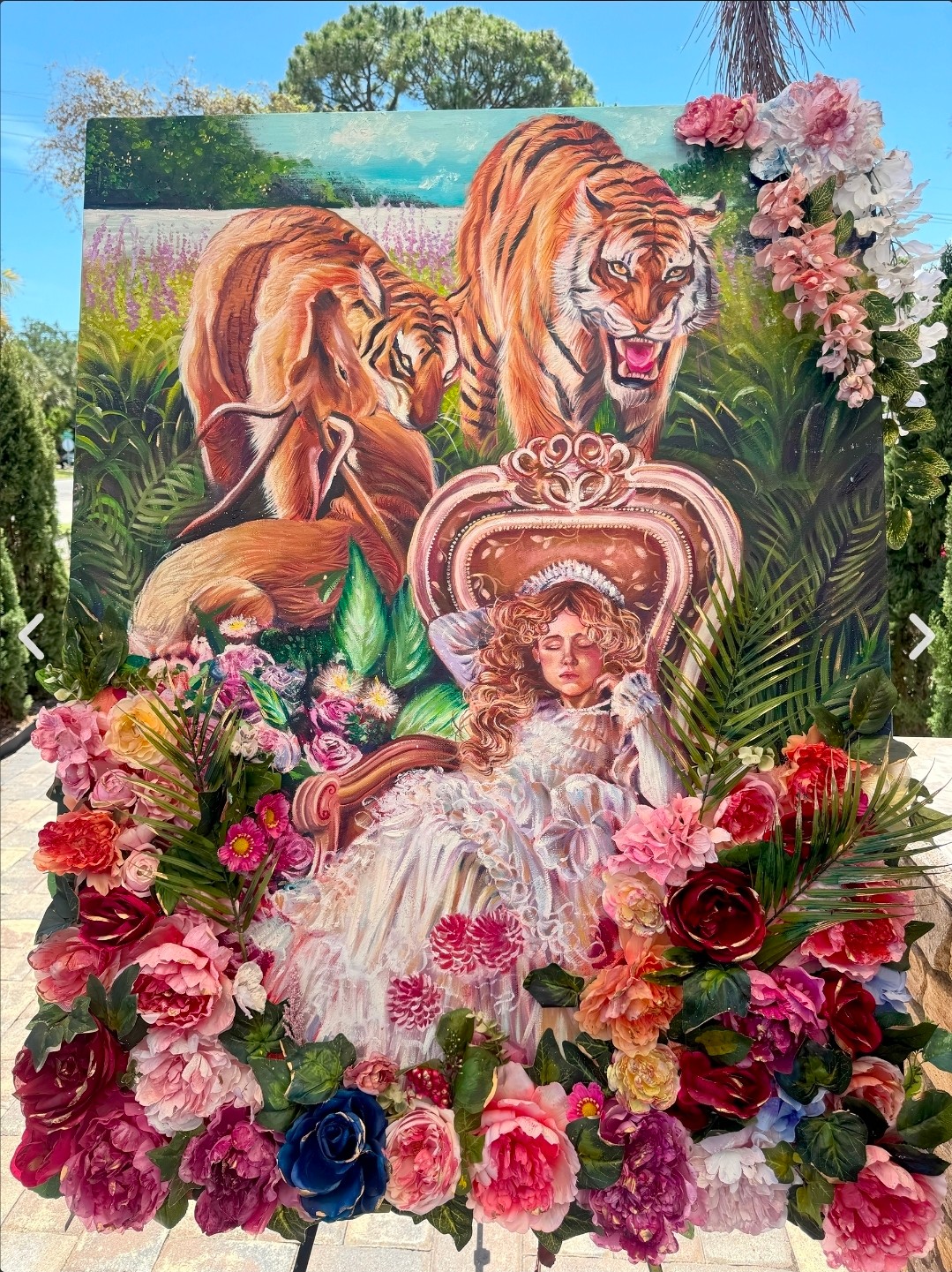 A painting of two lions and a lion cub surrounded by lush greenery, with a girl in a white dress sitting in front, surrounded by colorful flowers.