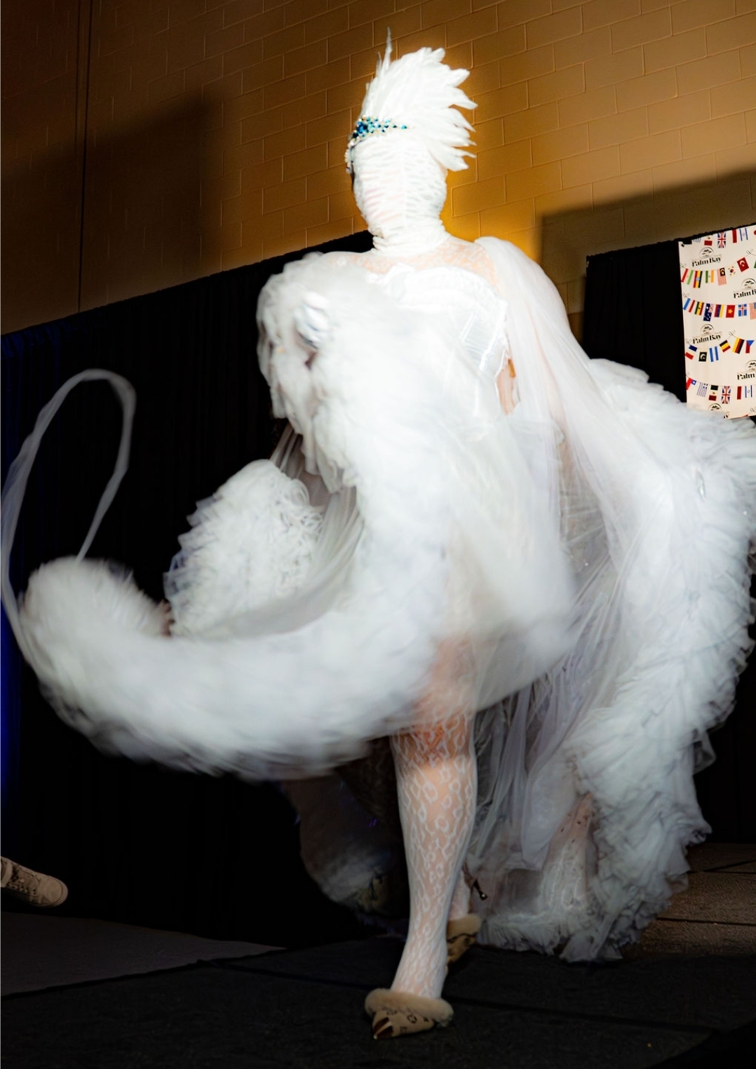 Person in elaborate white costume with feathers and flowing fabric, standing indoors near a black wall and a screen.