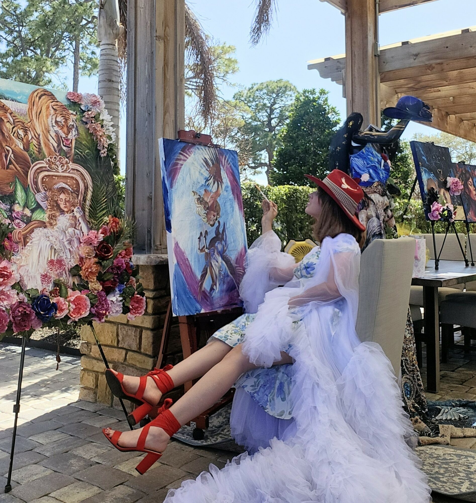 Child in a white dress and red shoes sitting on a chair, painting outdoors with art supplies and paintings nearby.