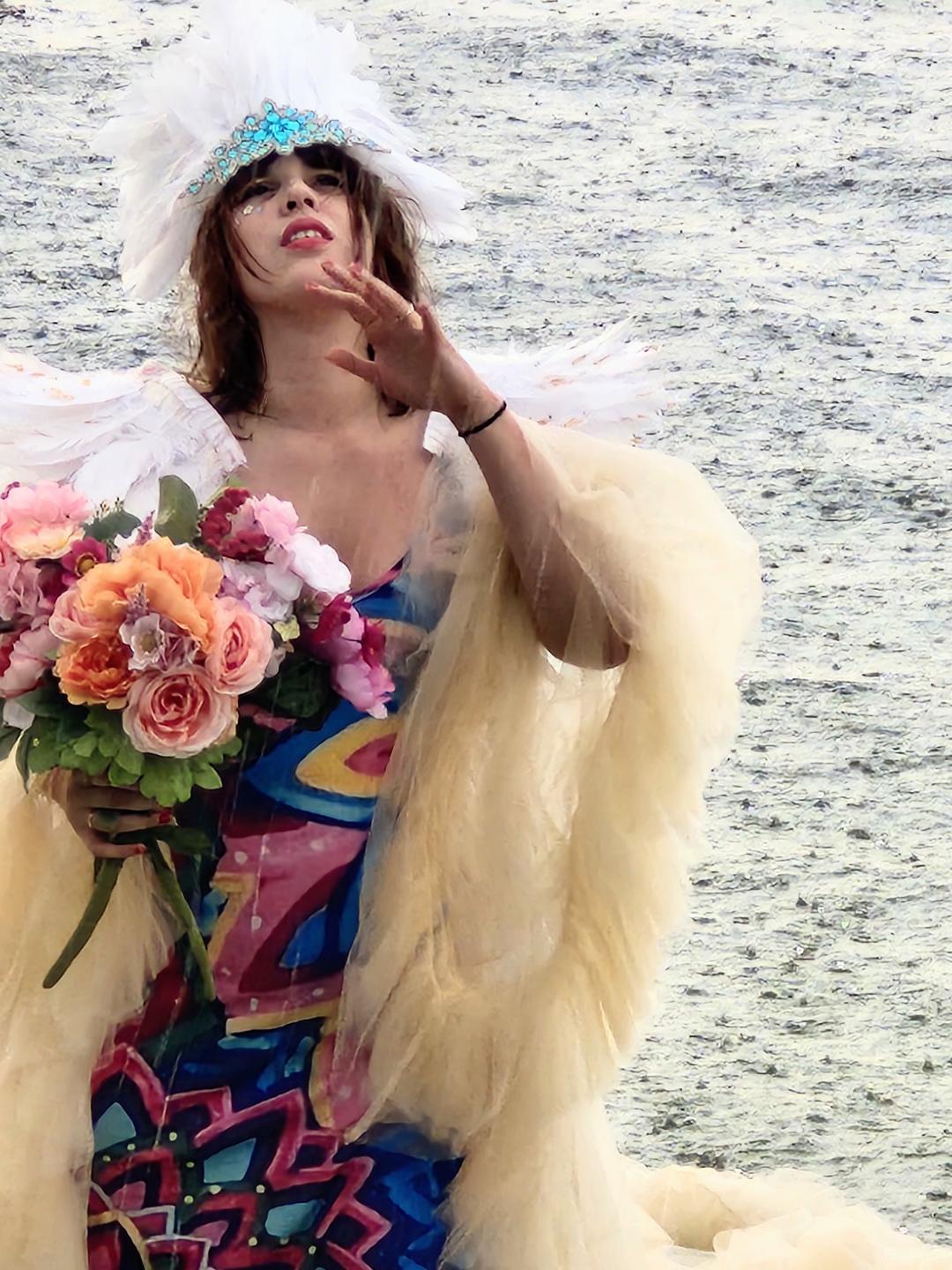 Woman in colorful dress and feathered hat holding a bouquet of flowers near water, with a yellow fur coat draped over her arm.