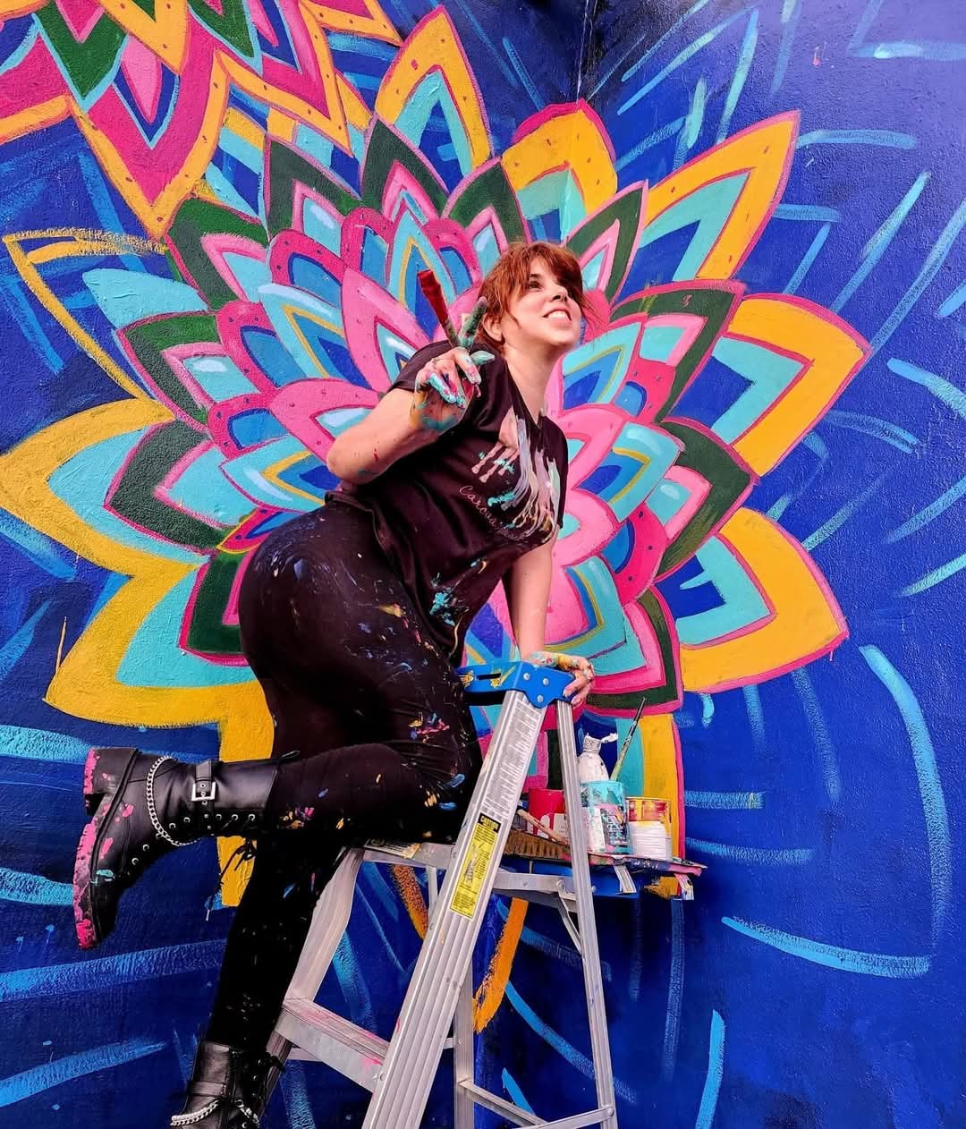 Person on ladder painting a colorful mandala mural on a wall, with vibrant patterns and blue background.
