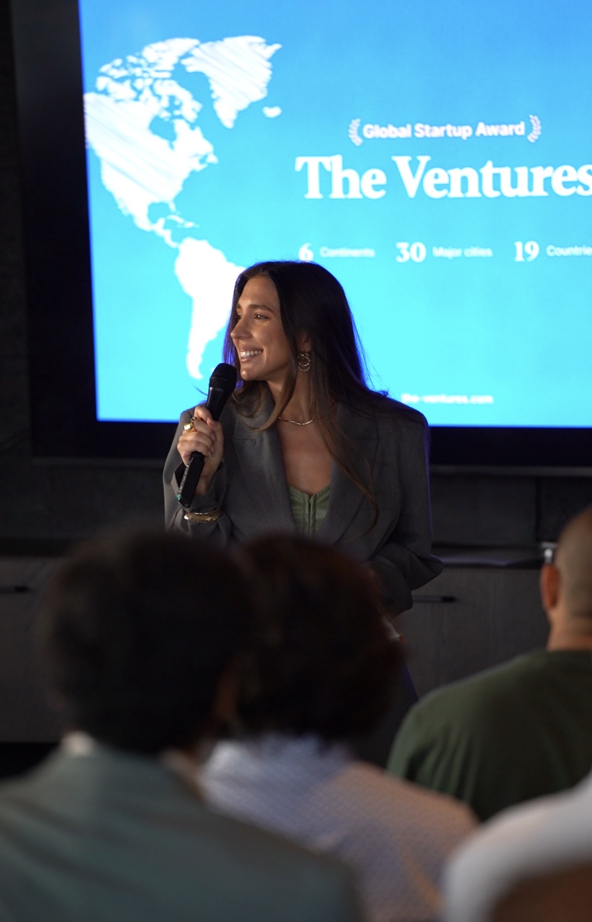 Woman speaking into microphone in front of a presentation screen with a world map and text, audience visible in foreground.