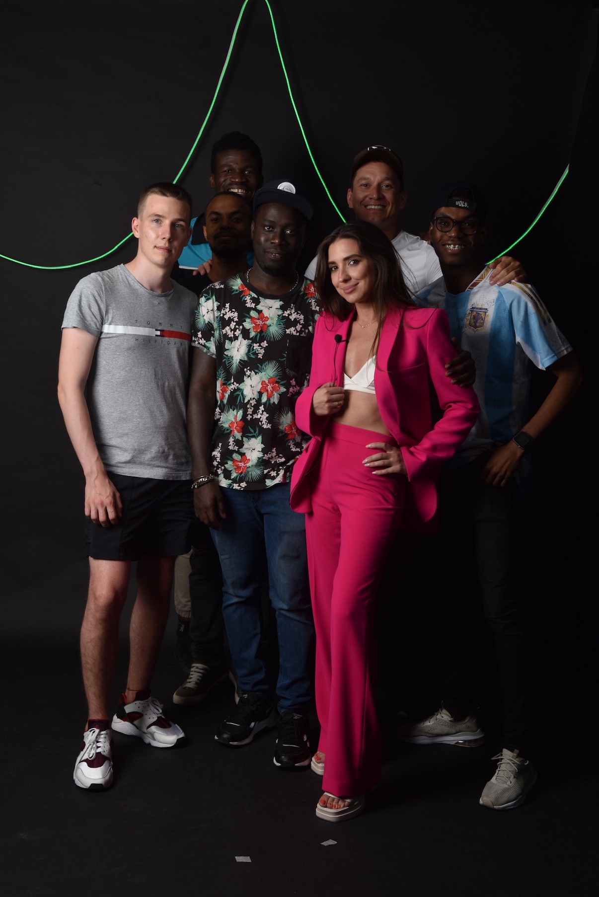 Group of six people standing in front of a black background with green lines, one woman in pink suit in front.