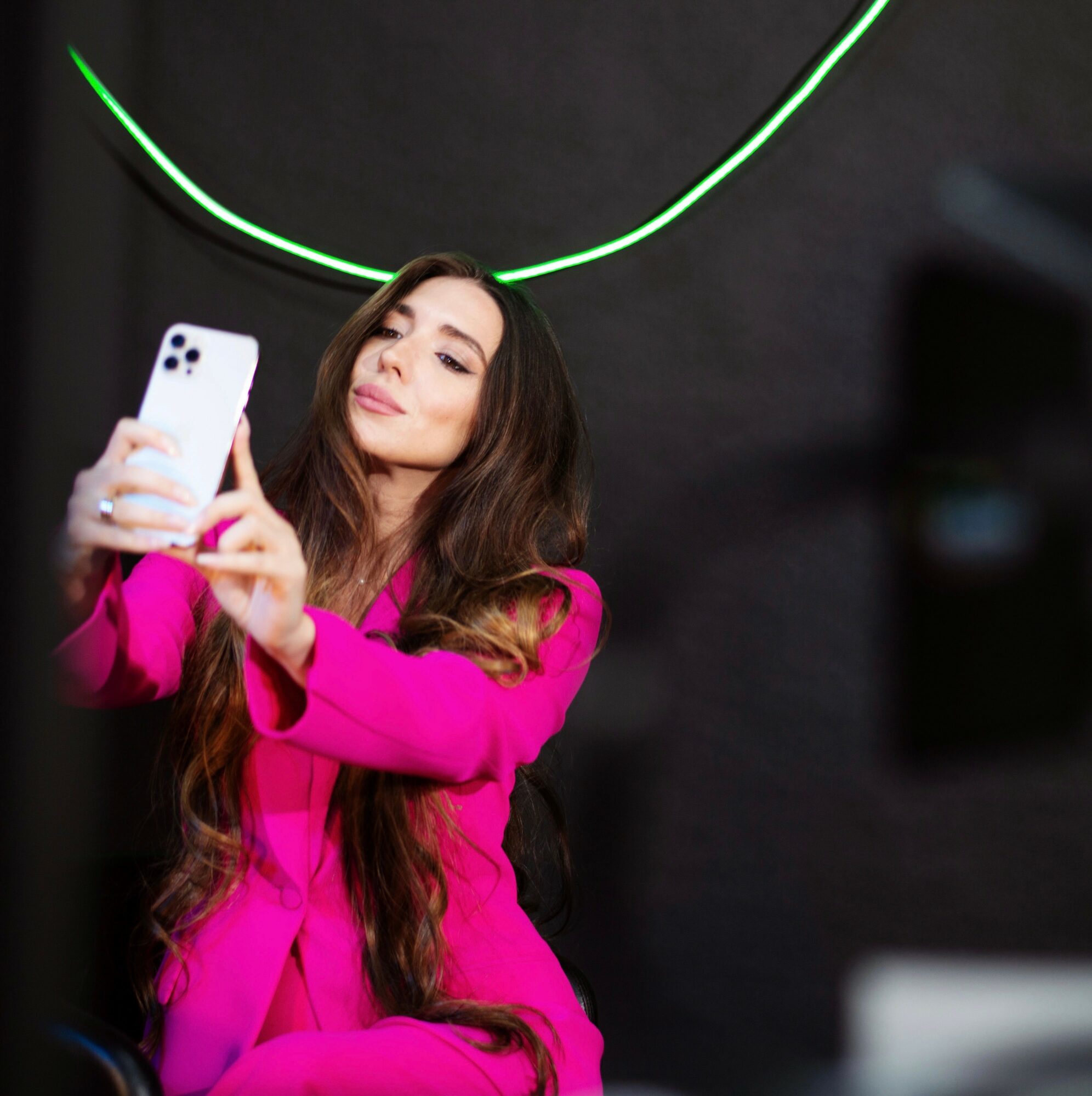 Woman with long hair in a pink outfit taking a selfie with a smartphone, dark background with green neon light arc.
