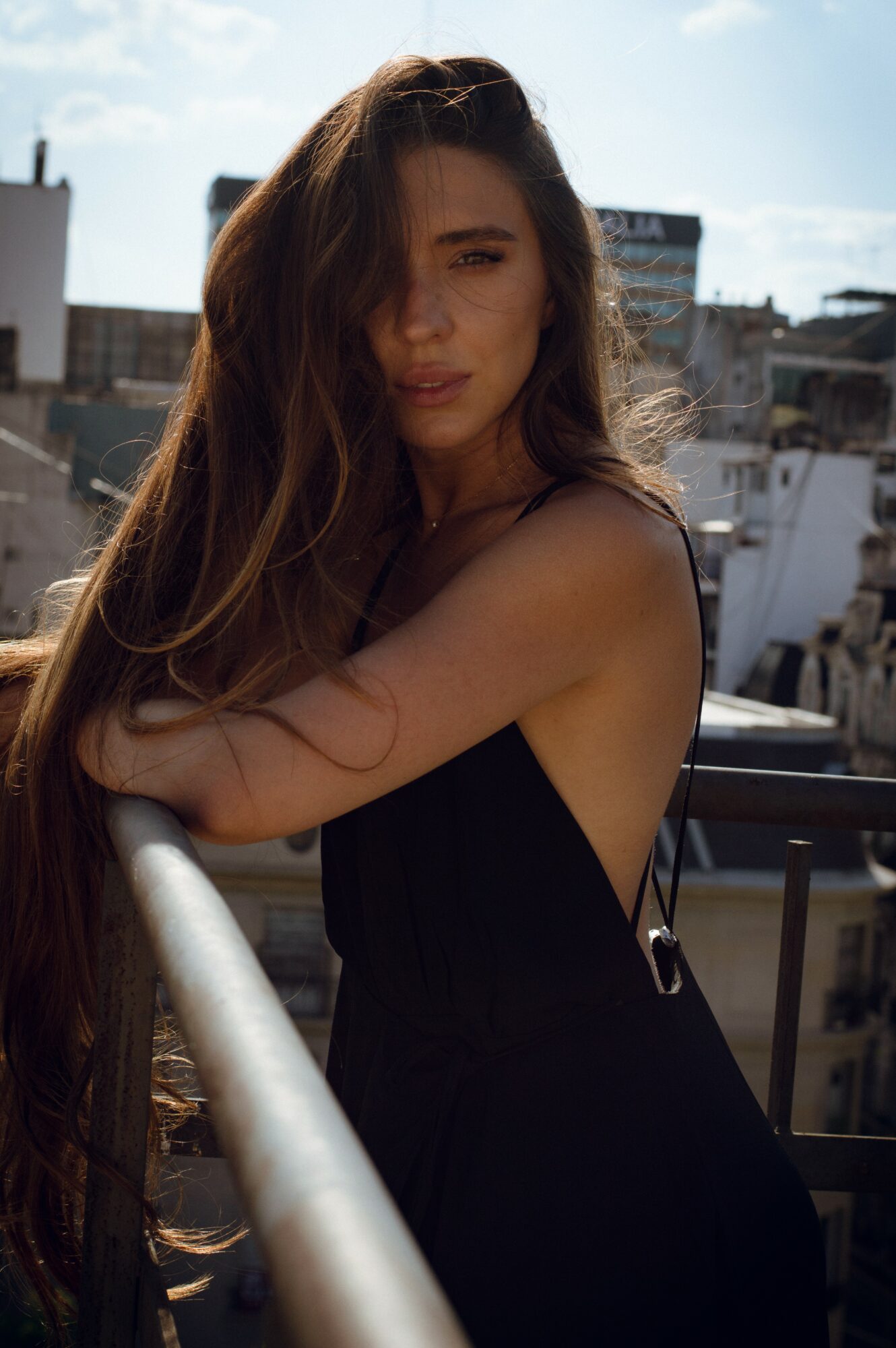 Young woman with long hair leaning on a balcony railing outdoors, cityscape background, sunlight, casual black outfit.