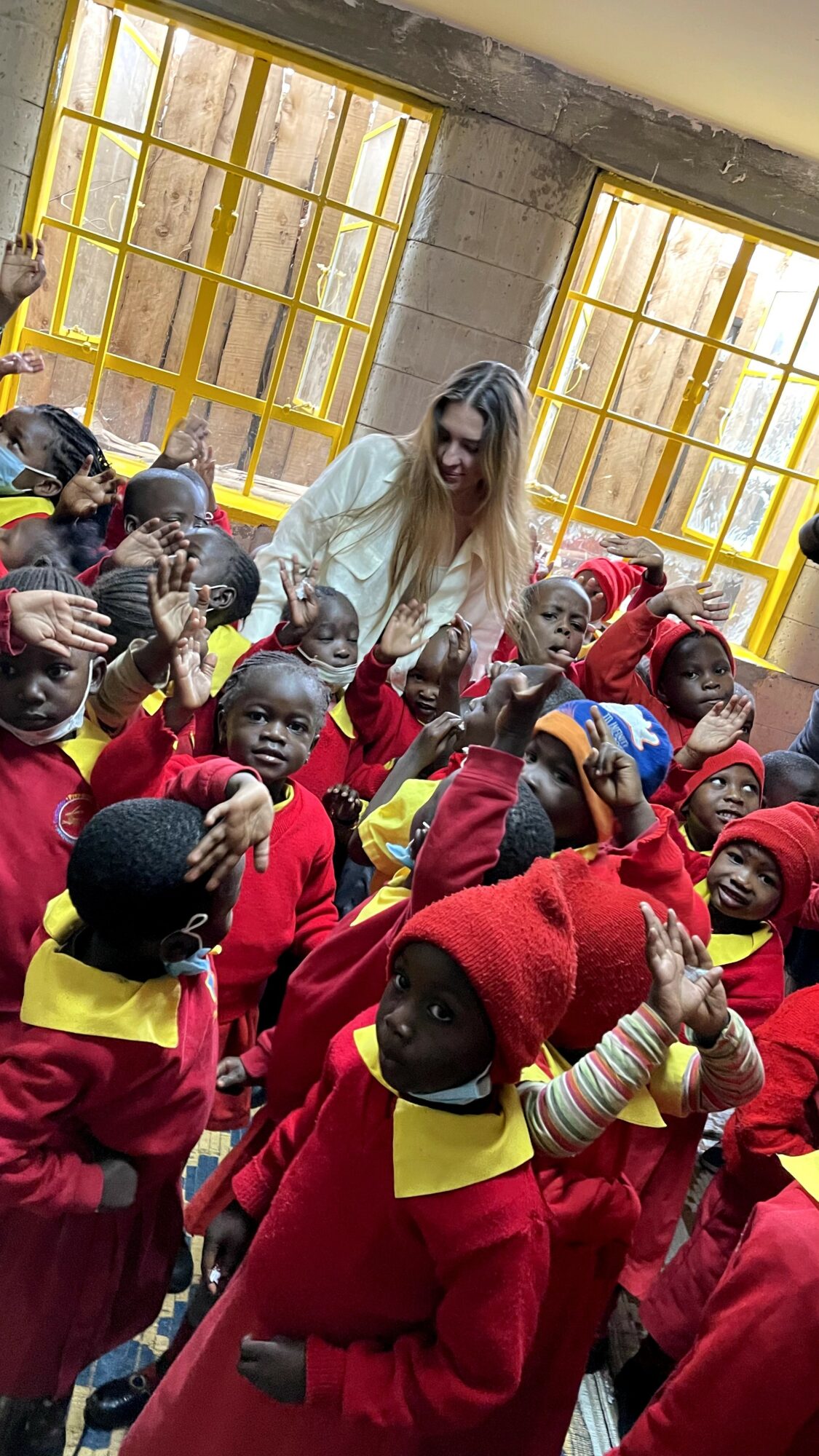 Group of children in red and yellow uniforms with a woman in a white jacket inside a room with large yellow windows.