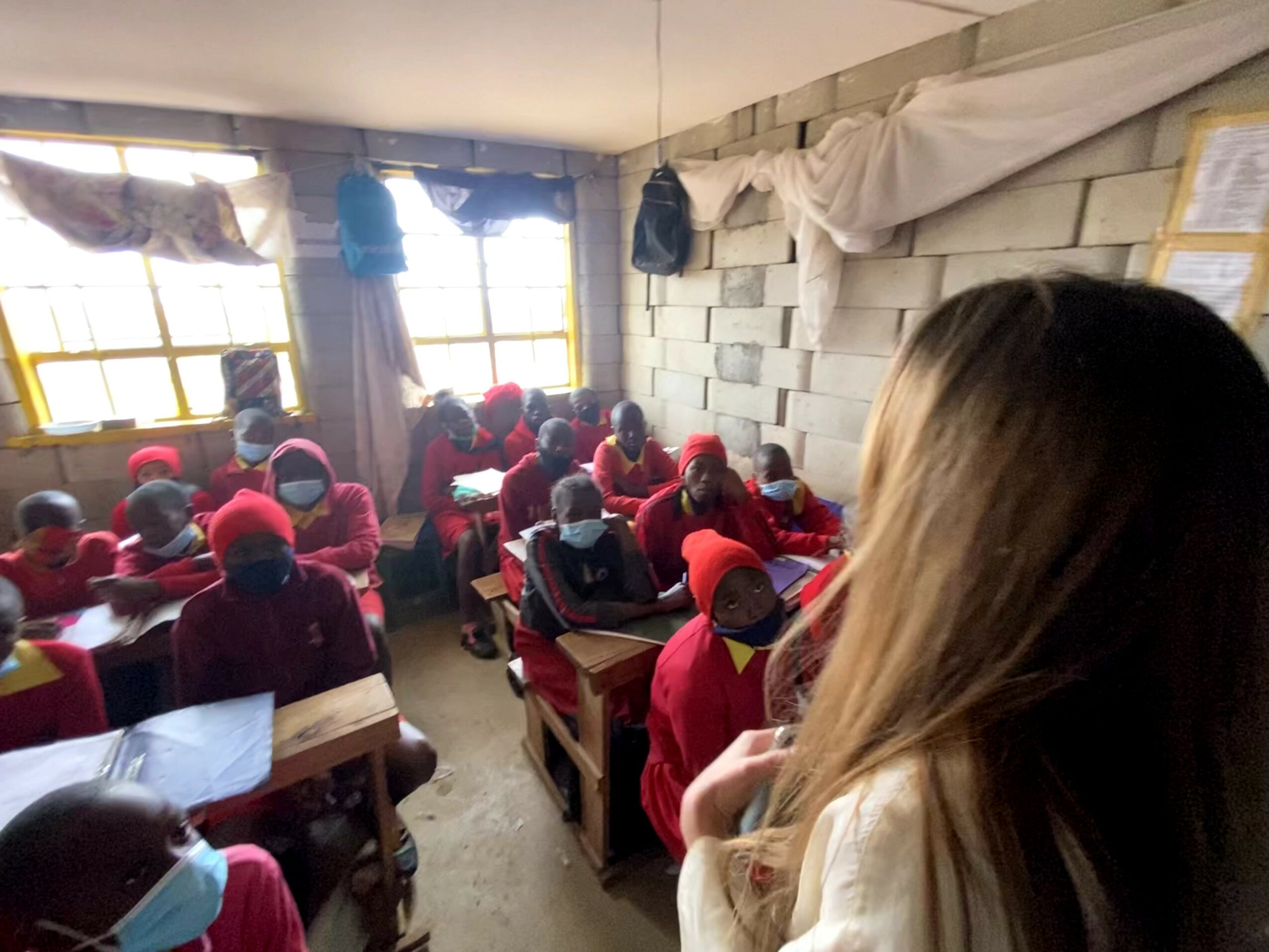Classroom with children wearing red uniforms and hats, sitting at desks, and a woman with long hair observing.