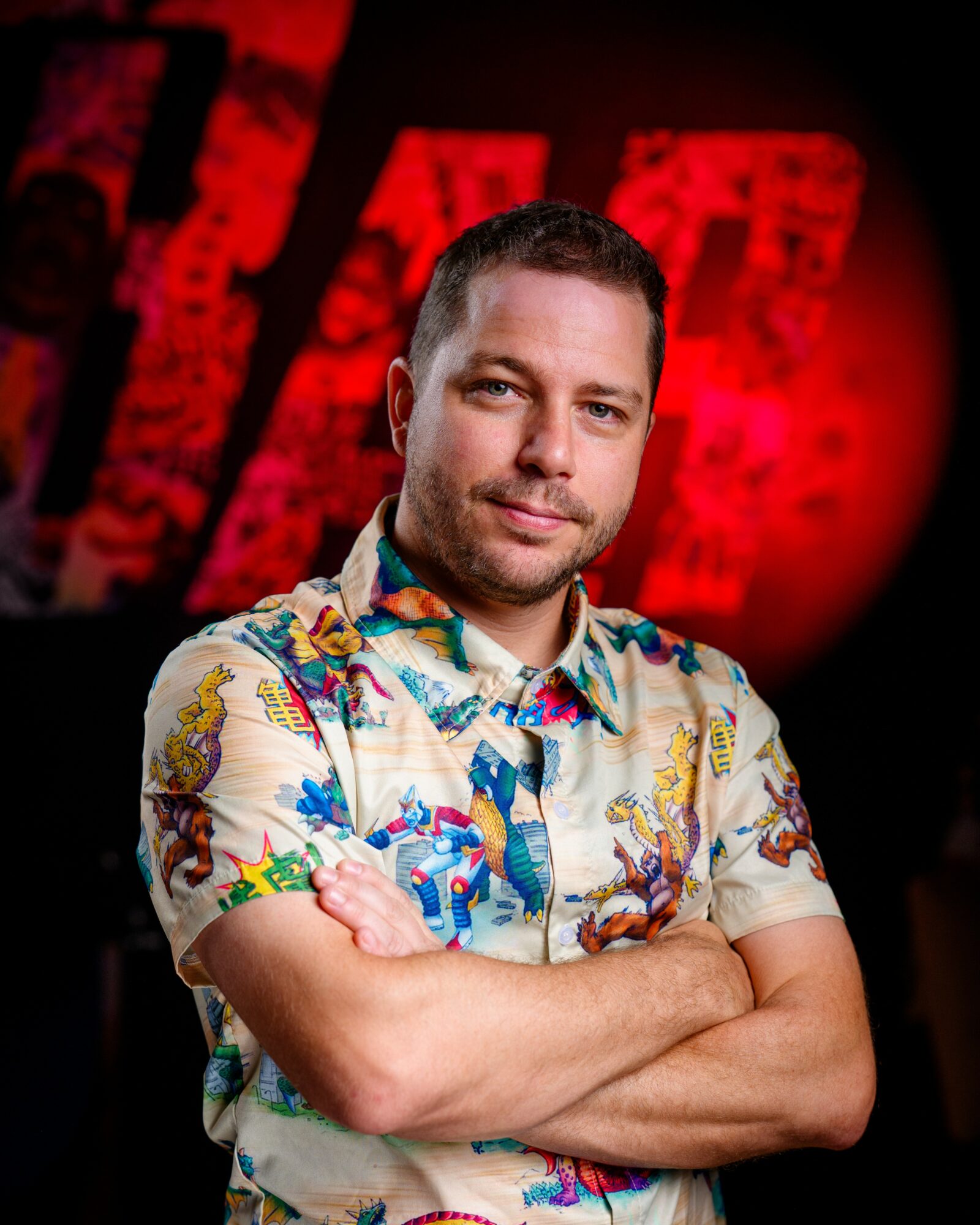 Man with crossed arms wearing a colorful floral shirt, standing in front of a red and black background.