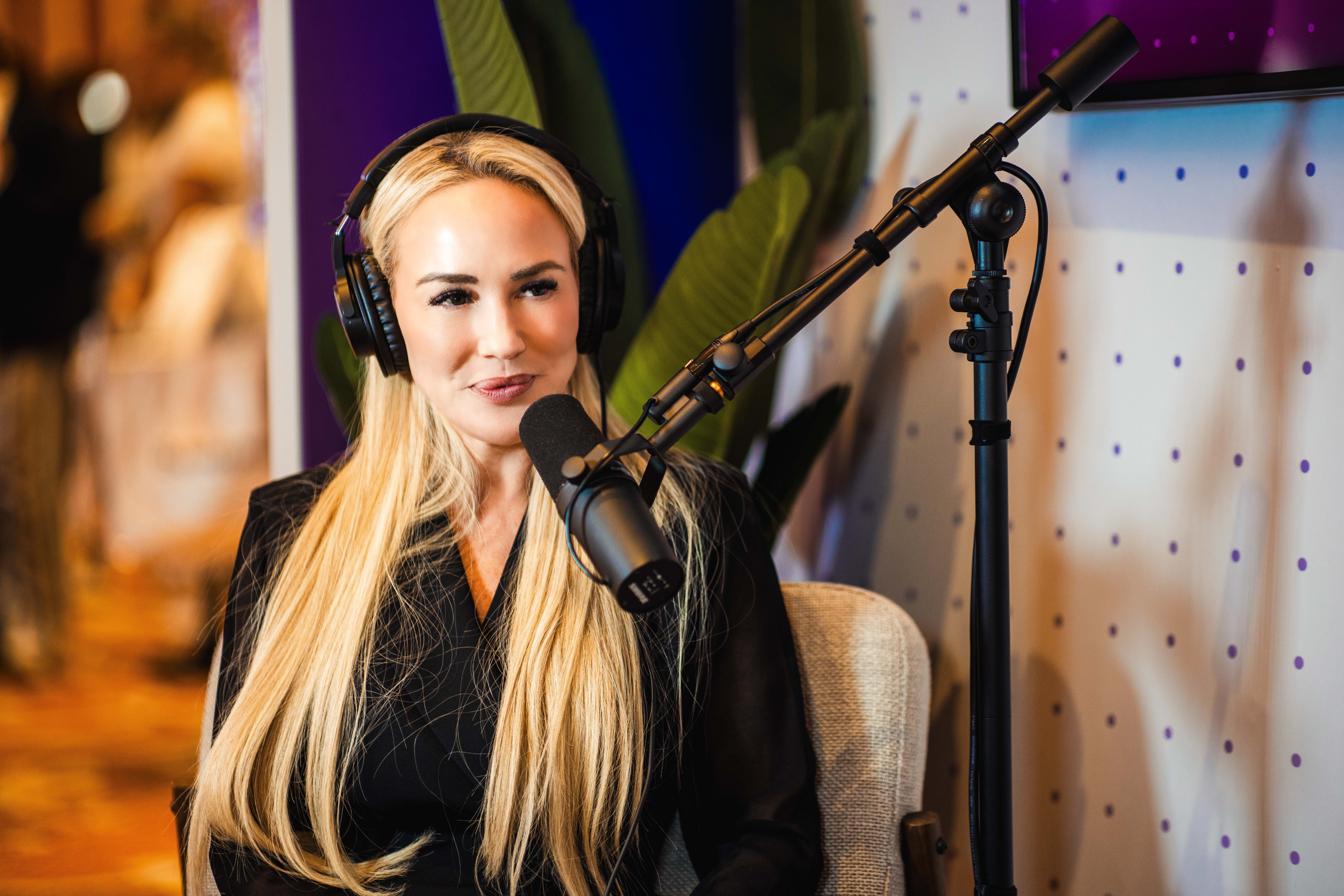 Woman with long blonde hair wearing headphones speaking into a microphone in a recording studio.