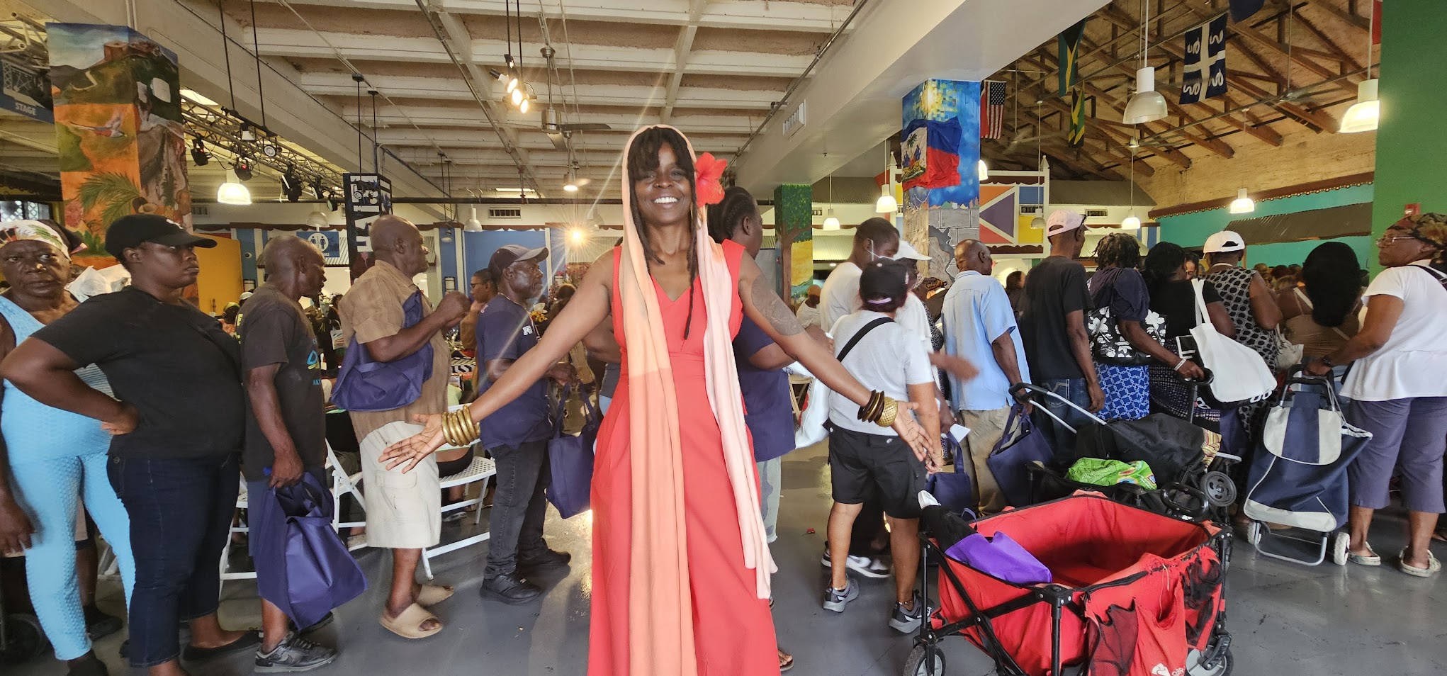 A woman in a pink dress stands with arms outstretched in a crowded indoor event space filled with people and booths.