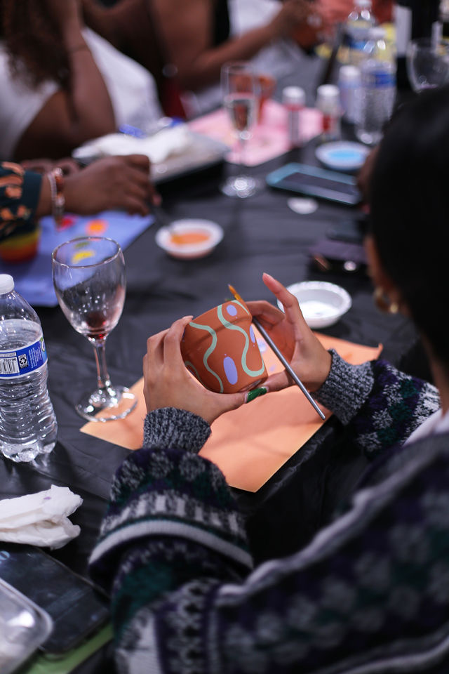 Person holding a decorated ceramic mug with a black straw, surrounded by drinks and tableware, at a gathering.
