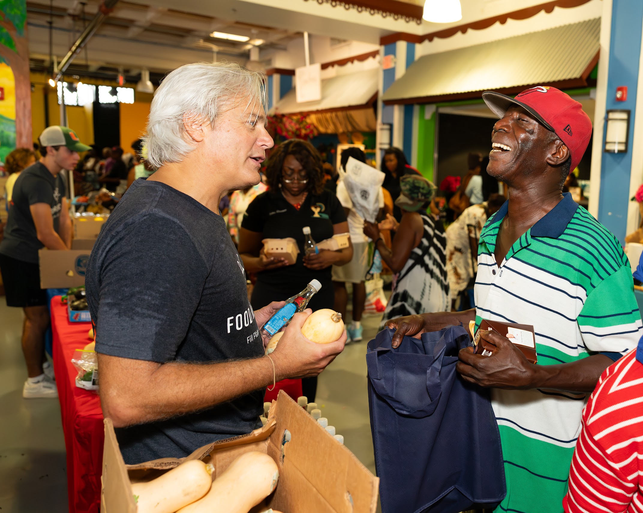 Two men smiling and talking at a market stall with people in the background.