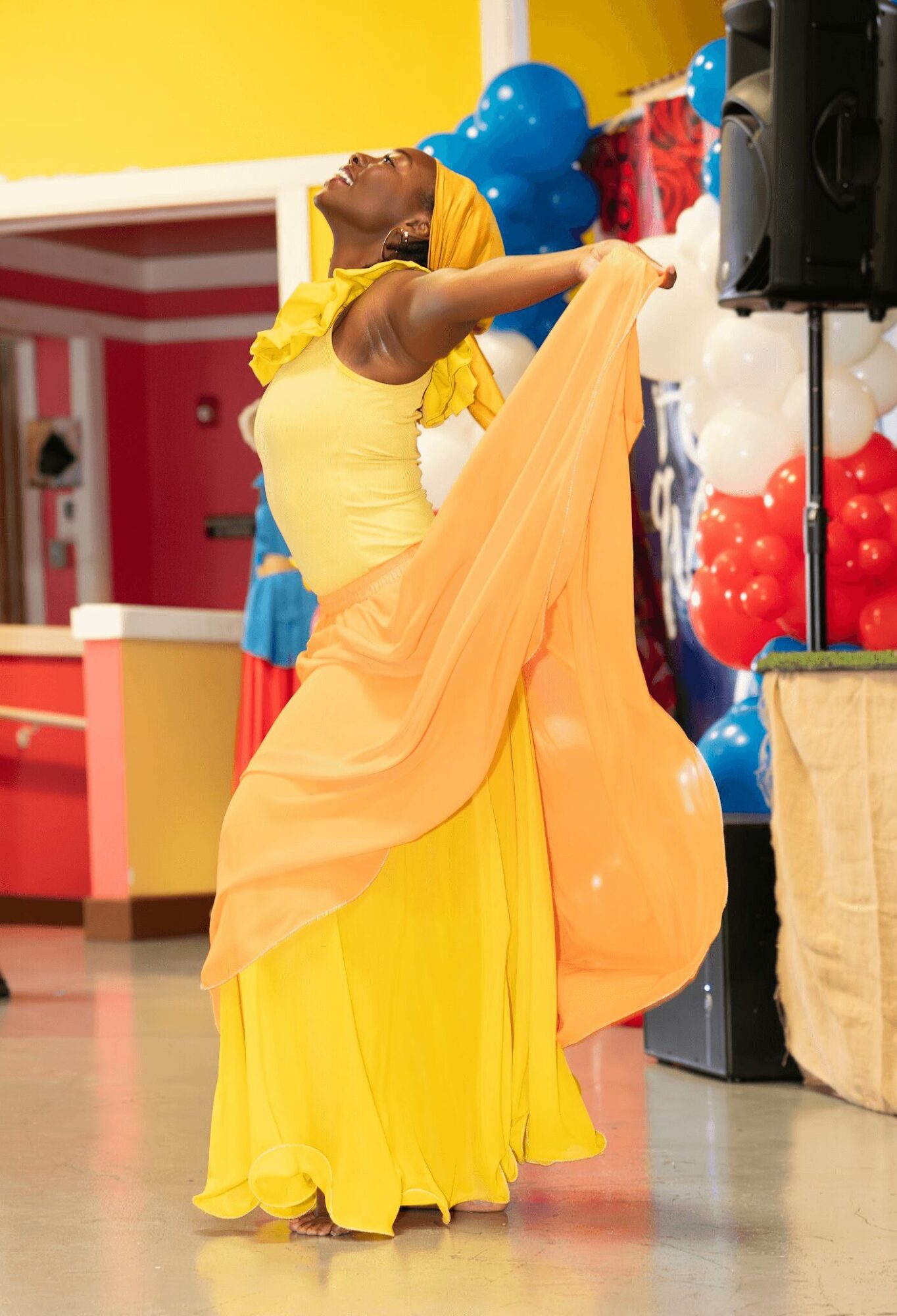 Person dancing in yellow and orange dress with balloons and decorations in background.