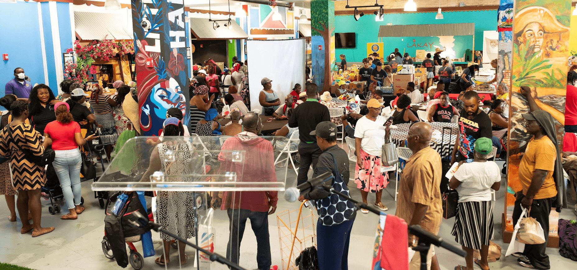 Crowd gathered in an indoor event space with colorful decorations and a stage, people standing and walking around.