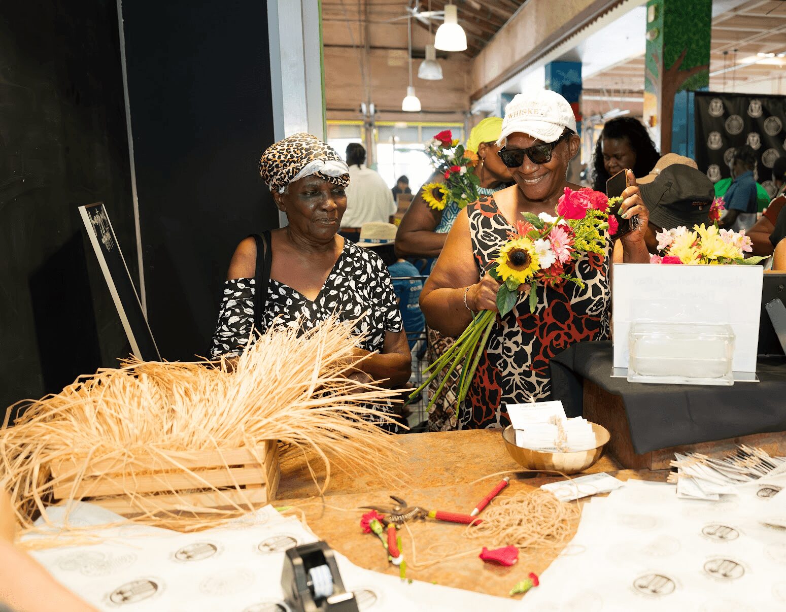 Two women at a market stall with flowers and wheat, one holding a bouquet, in a busy indoor setting.