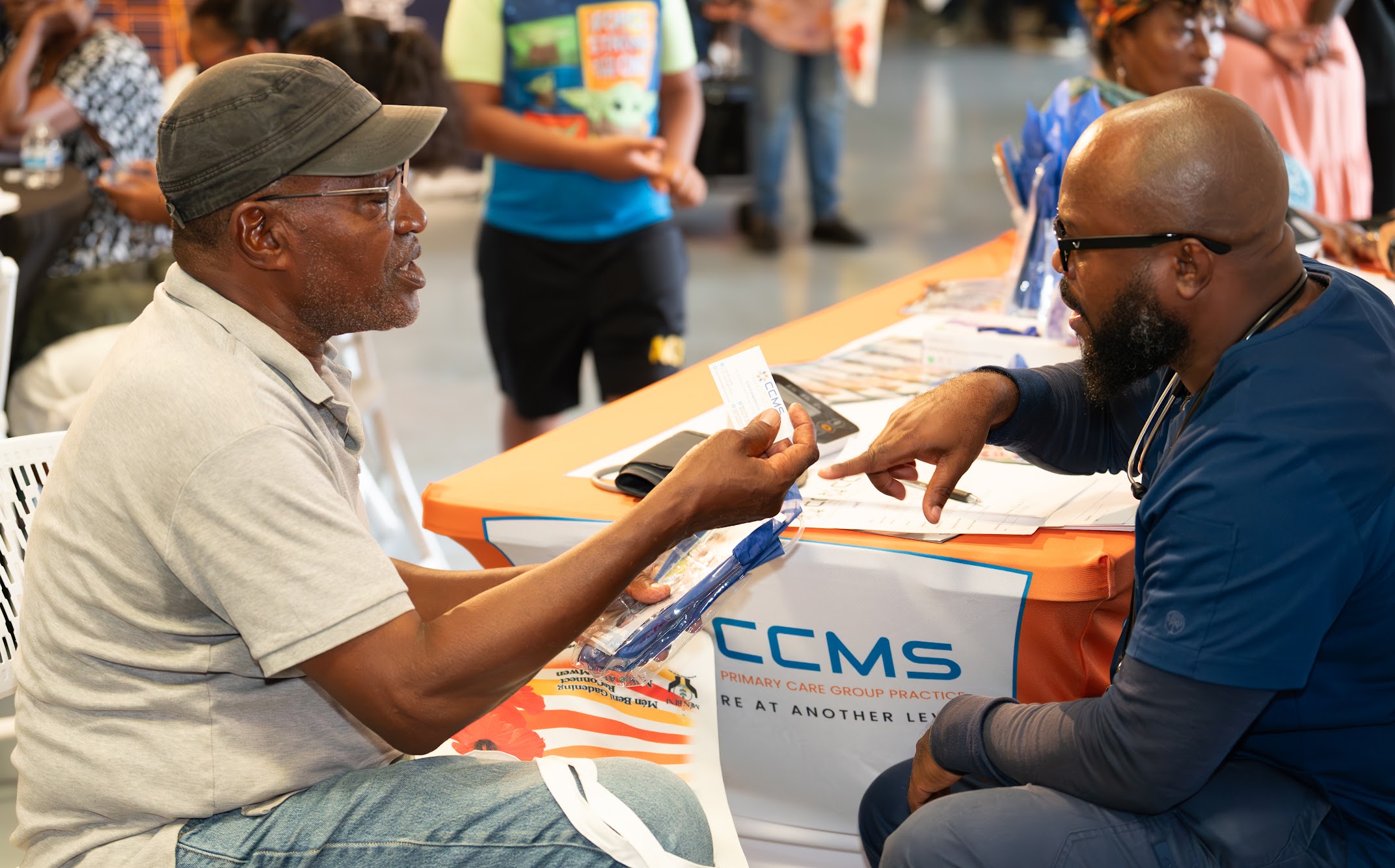 Two men sit at a table engaged in conversation, with people and a table in the background.