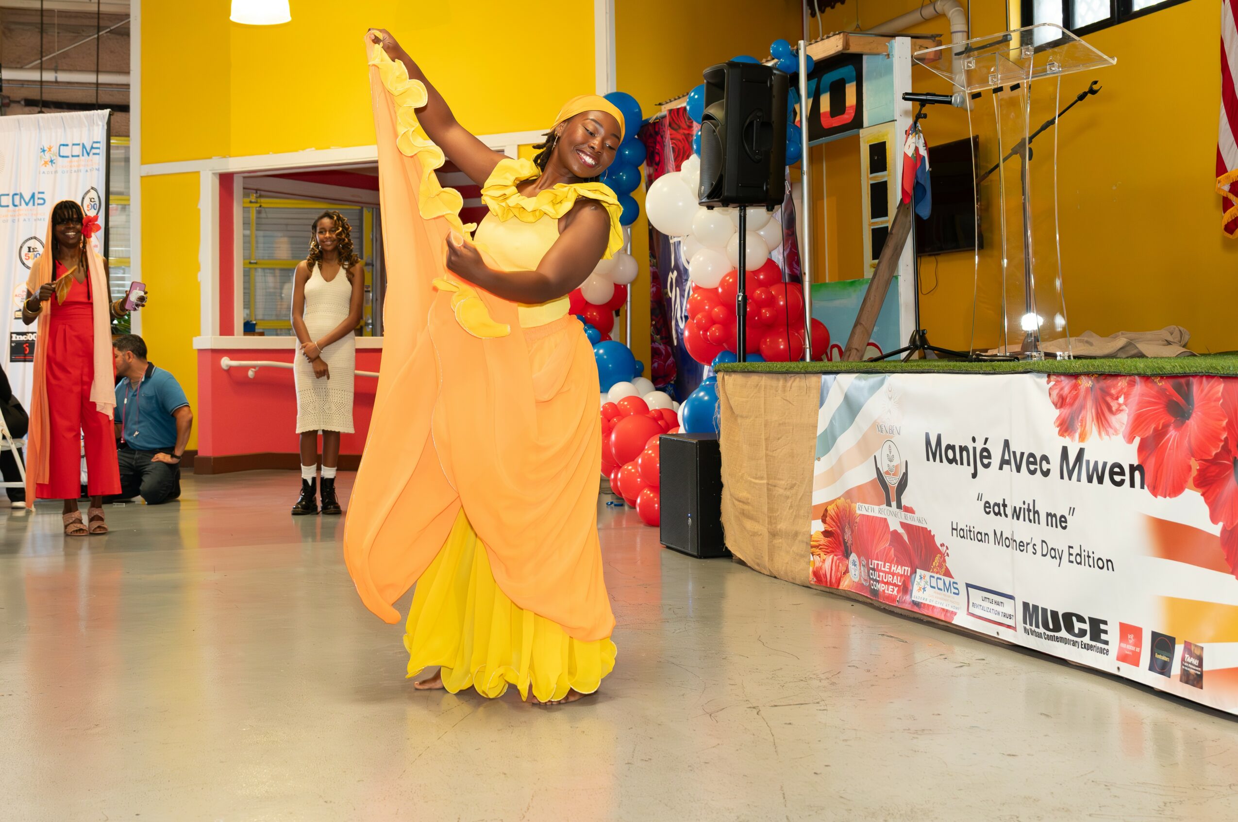 Person in colorful dress dancing with arms raised in a decorated indoor space, balloons and a banner in the background.