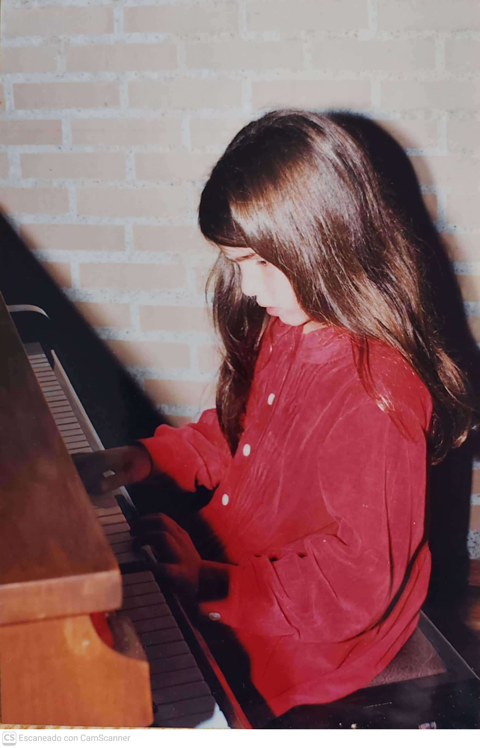 Girl with long brown hair playing piano, wearing a red shirt, against a brick wall background.