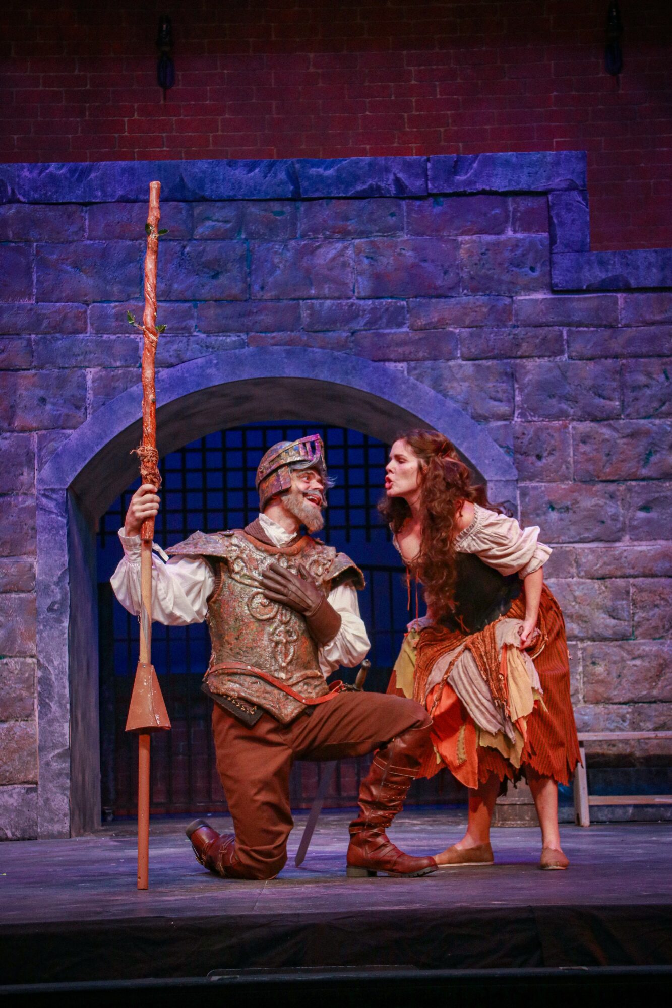 Two actors in medieval costumes perform on stage in front of a stone wall backdrop, one holding a staff, the other kneeling.