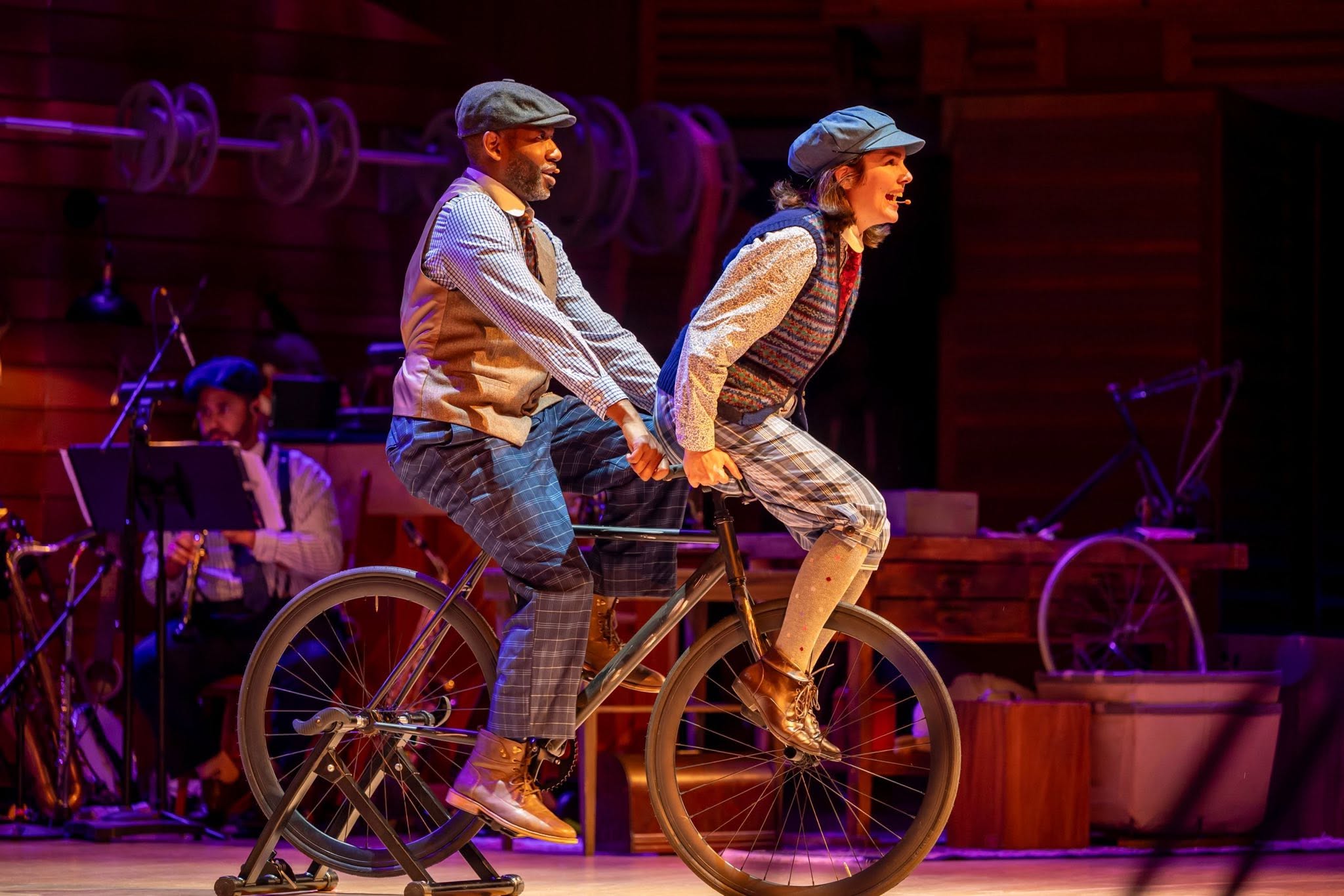Two people riding a tandem bicycle on stage with musicians in the background.