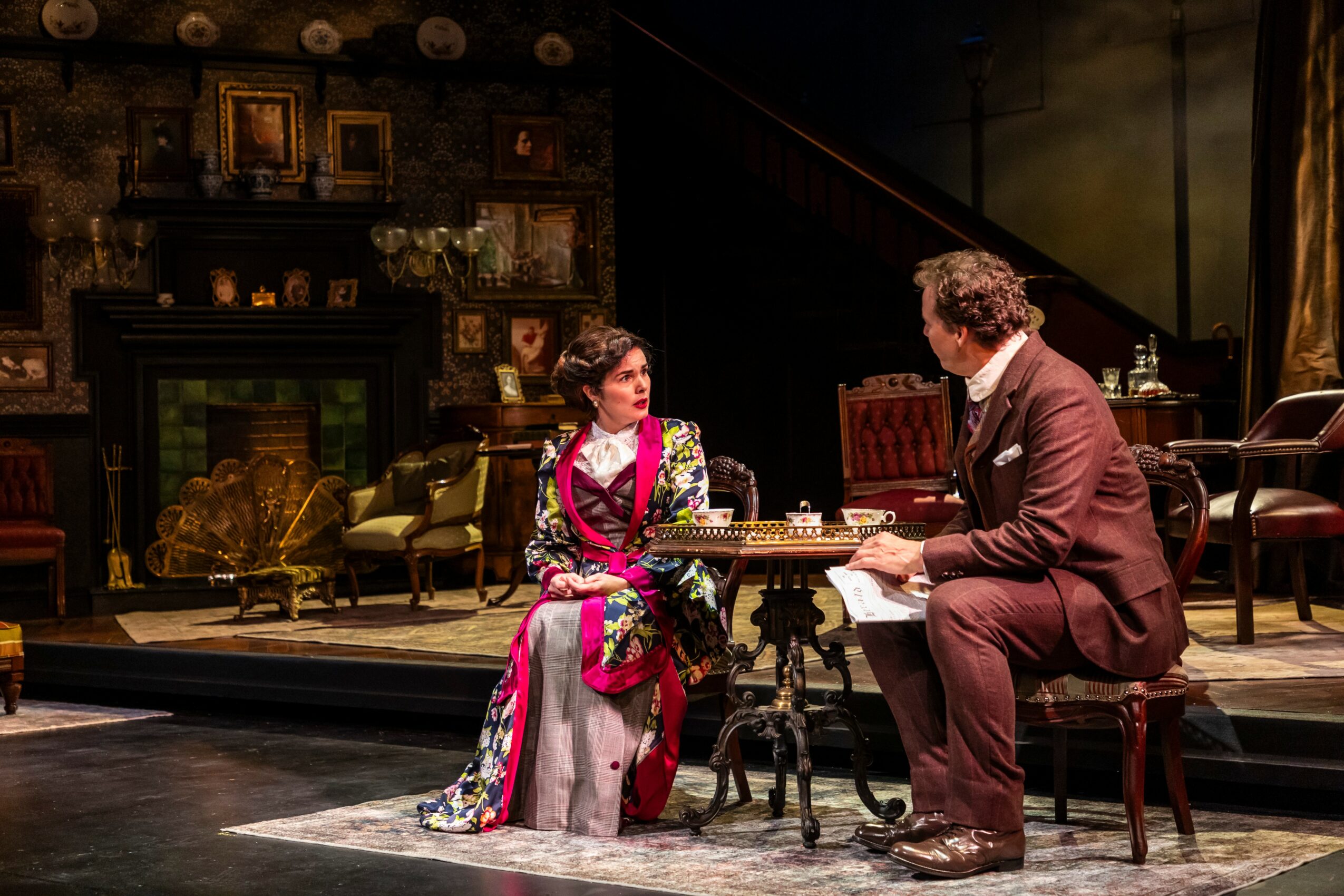Two actors sit at a table on stage, engaged in conversation, with a detailed set including furniture and decorations behind them.
