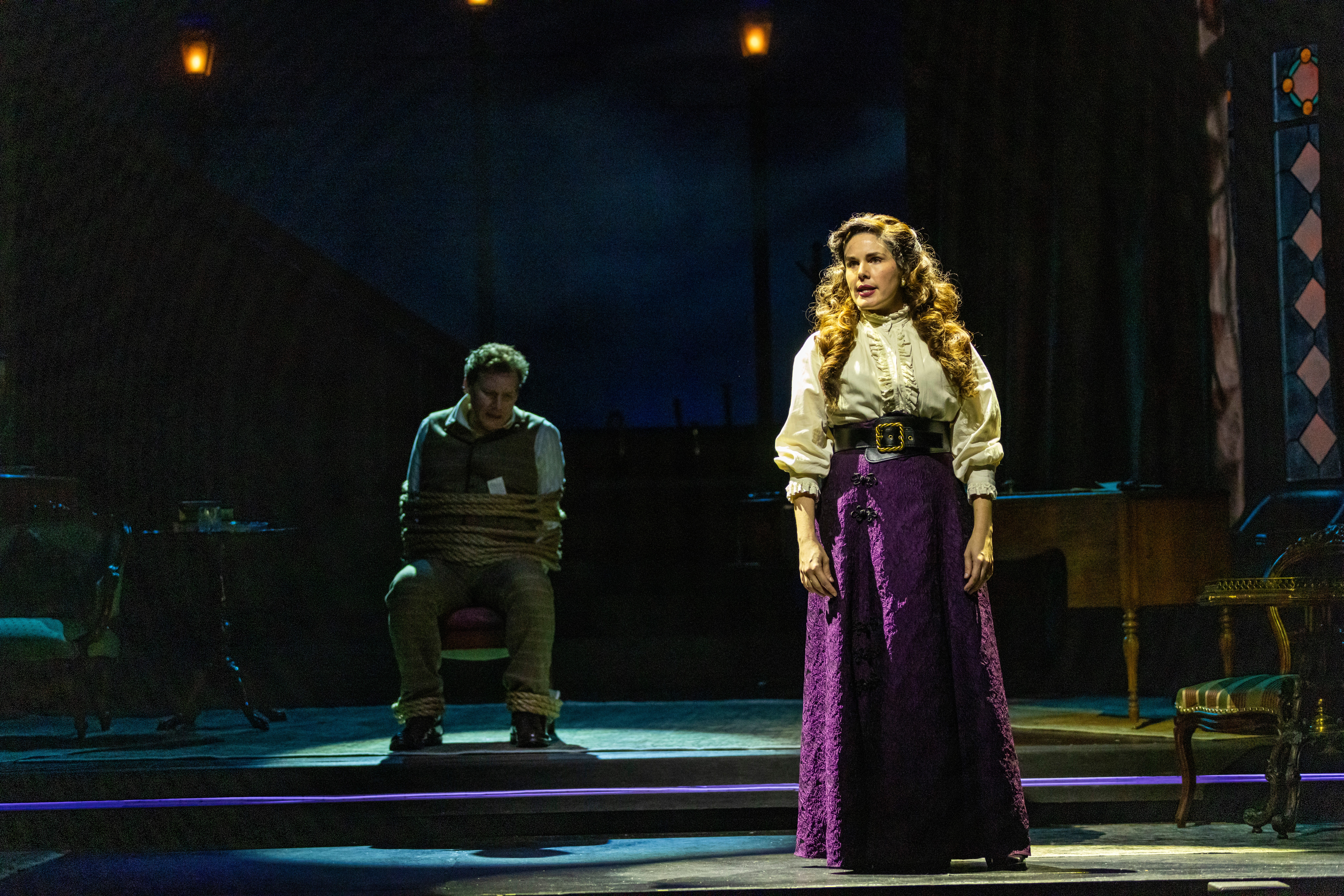 A woman with long, curly hair stands on stage in a purple skirt and white blouse, with a man sitting behind her.