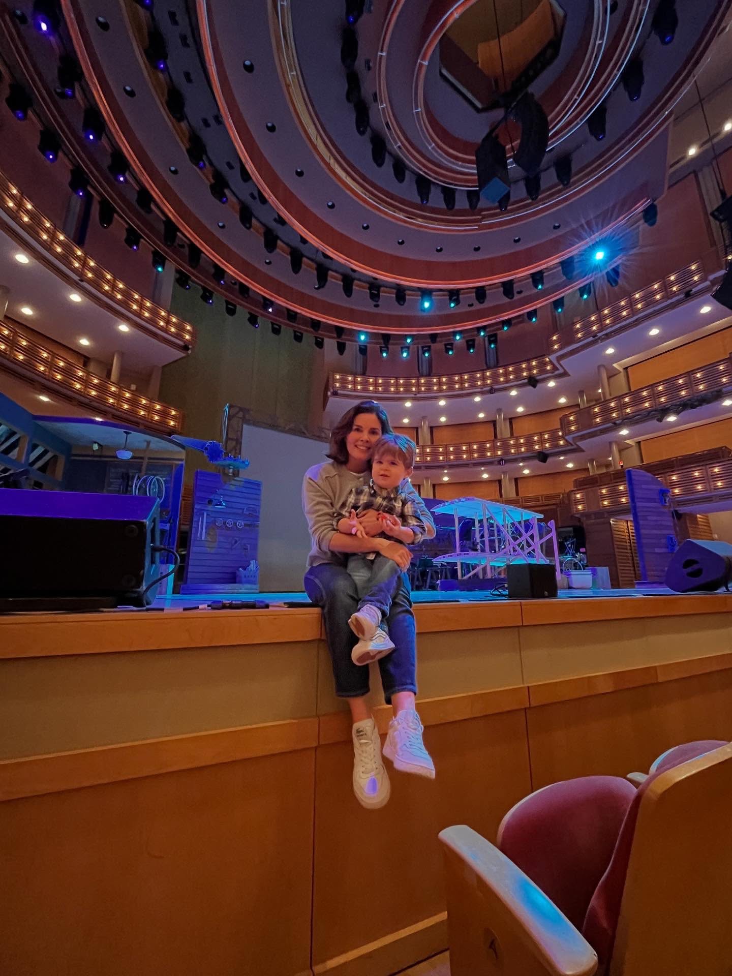 Two people sitting on a stage in a theater, with a large circular ceiling and balconies, stage lights, and empty seats.