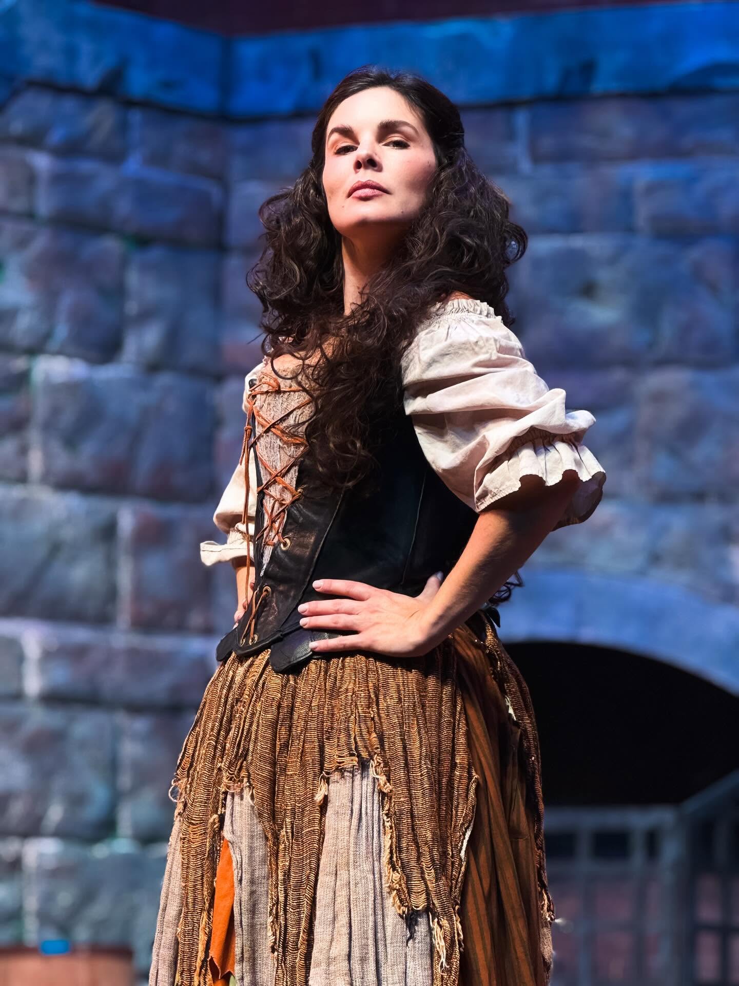 Woman with long curly hair in historical costume standing outdoors against stone wall, hands on hips, looking confident.