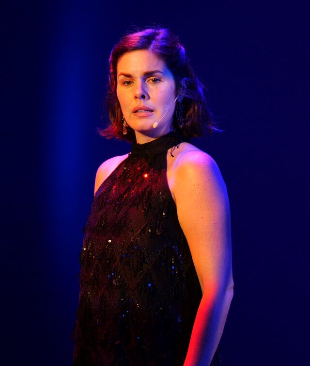 Woman with shoulder-length dark hair in a black, sparkly sleeveless dress, standing against a dark blue background.